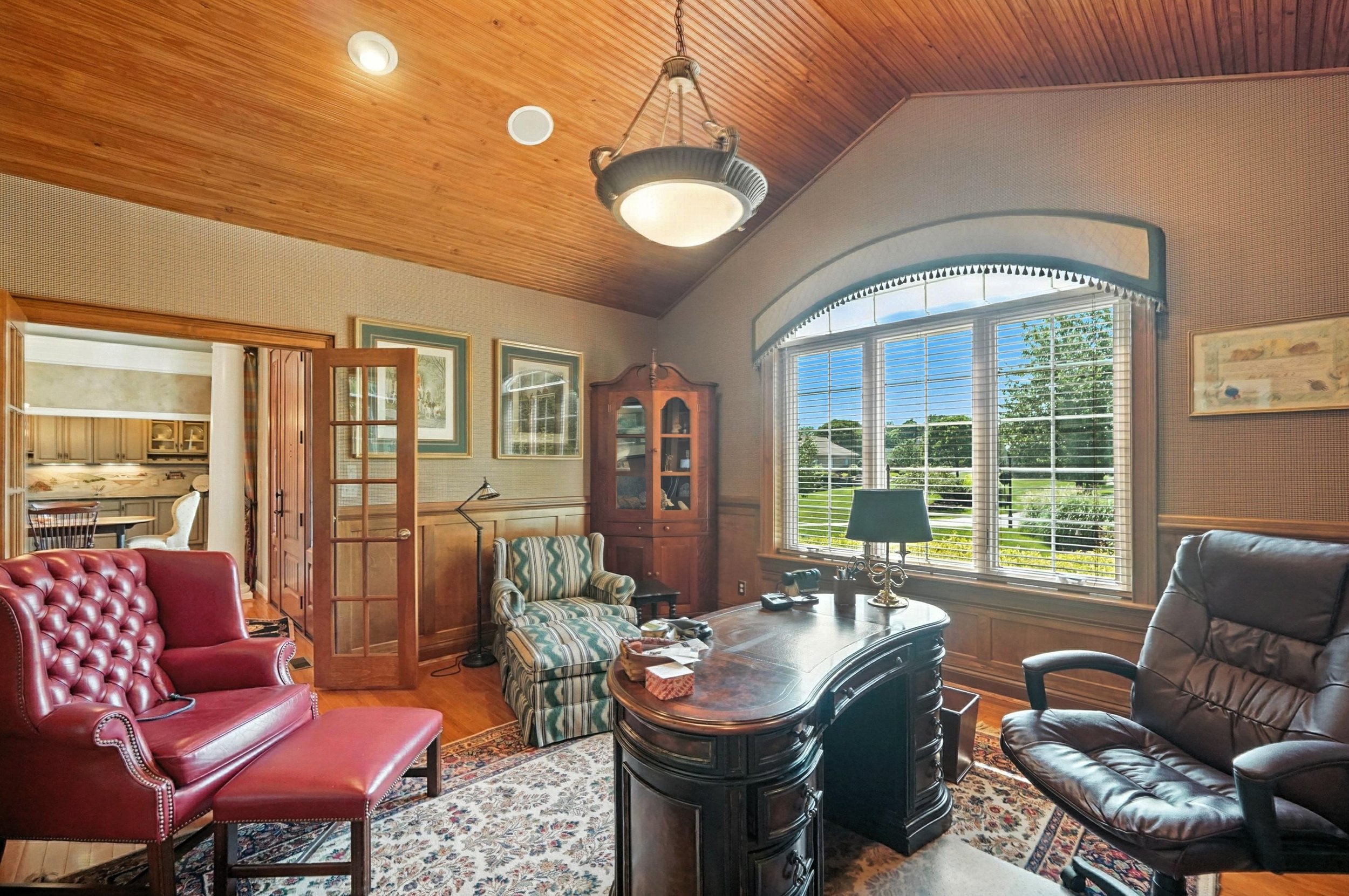 Living room with wooden ceiling, large window with blinds showing outdoor greenery, red leather armchair with matching ottoman, striped armchair, black desk, and a leather recliner chair.