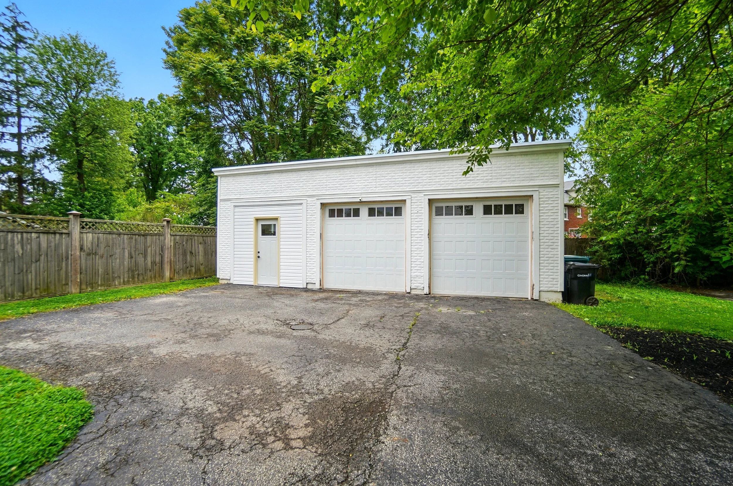 Backyard garage with two white doors, one small side door, on a cracked asphalt driveway, surrounded by green trees and grass.
