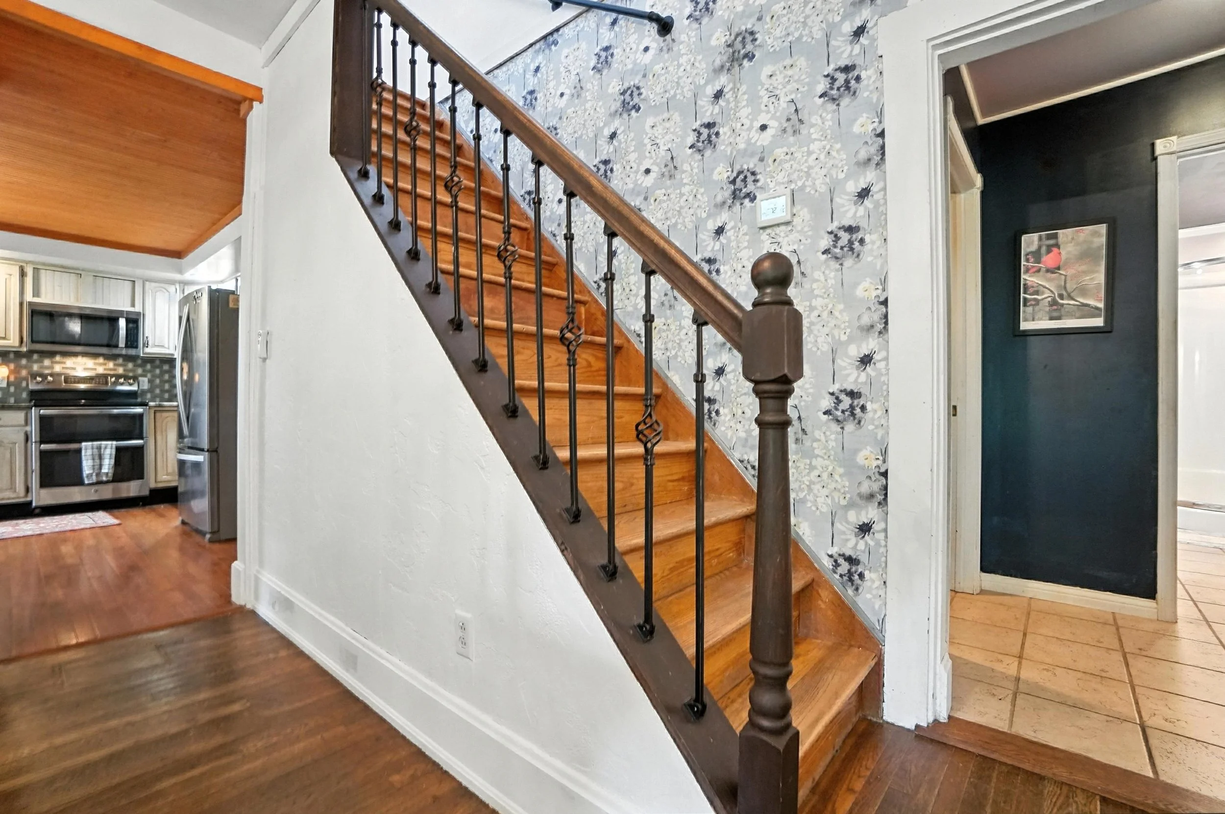 Interior of a house showing a staircase with wooden steps and a dark wood banister. The wall next to the staircase has floral wallpaper. To the left is a kitchen area with light cabinetry and a stainless steel refrigerator. To the right, a doorway wi