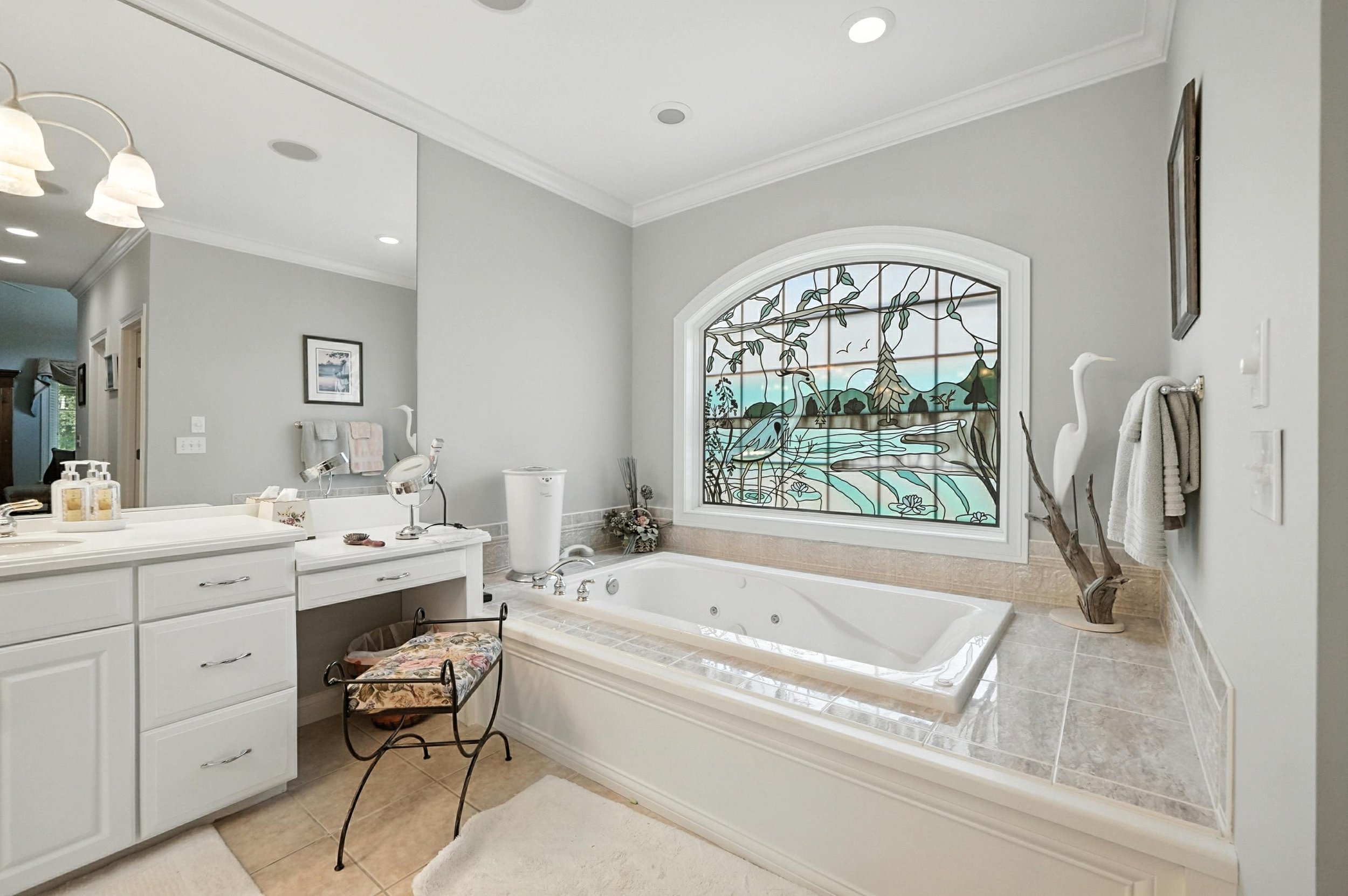 Bathroom with a large bathtub in front of a stained glass window depicting a nature scene with cranes, trees, and mountains. The bathroom has white cabinets, a vanity with a mirror, and decorative items including towels, a stylish chair, and a sculpt