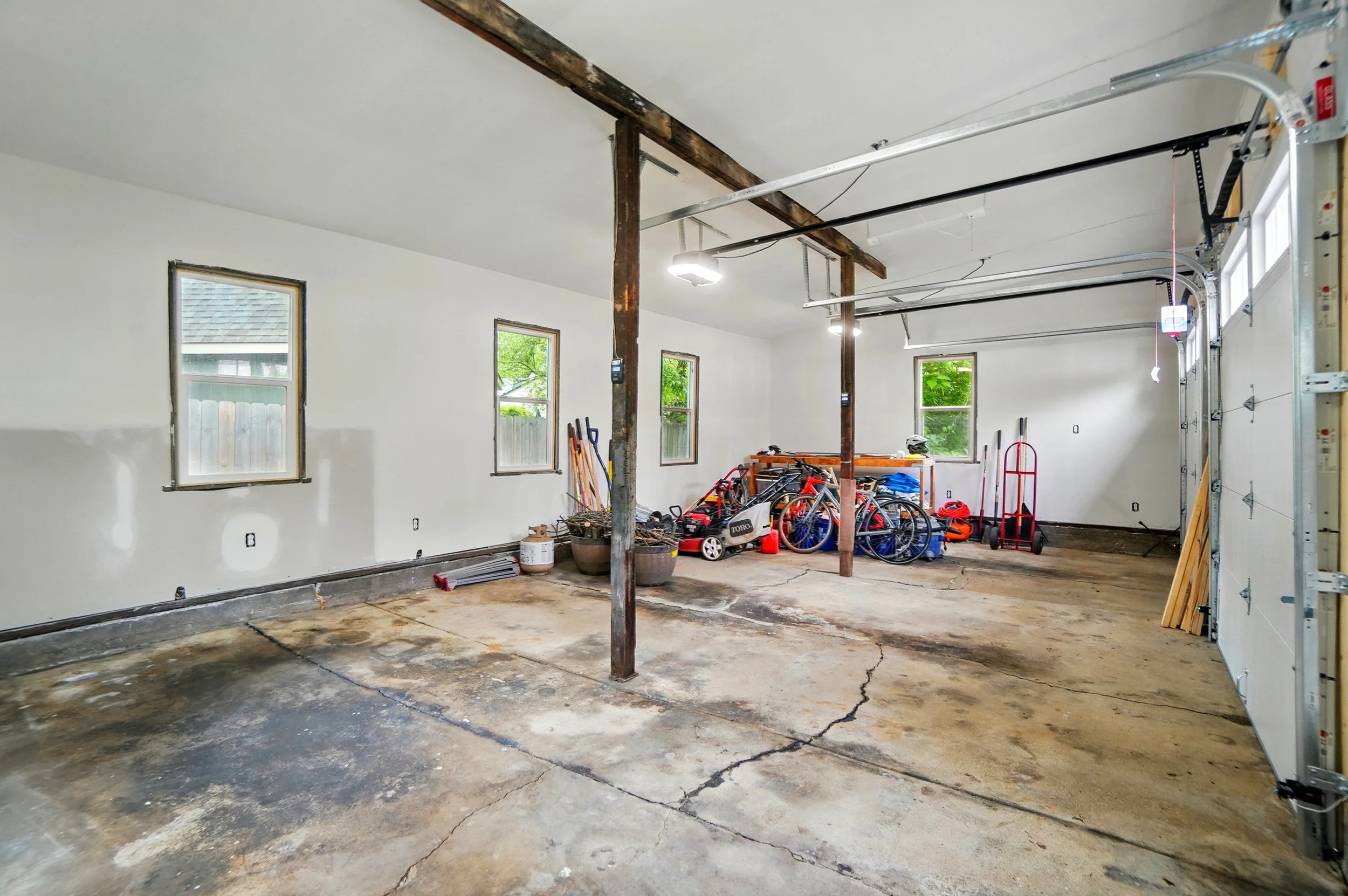 Empty garage with cracked concrete floor, white walls, three windows, and organized tools and bicycles near the back wall.