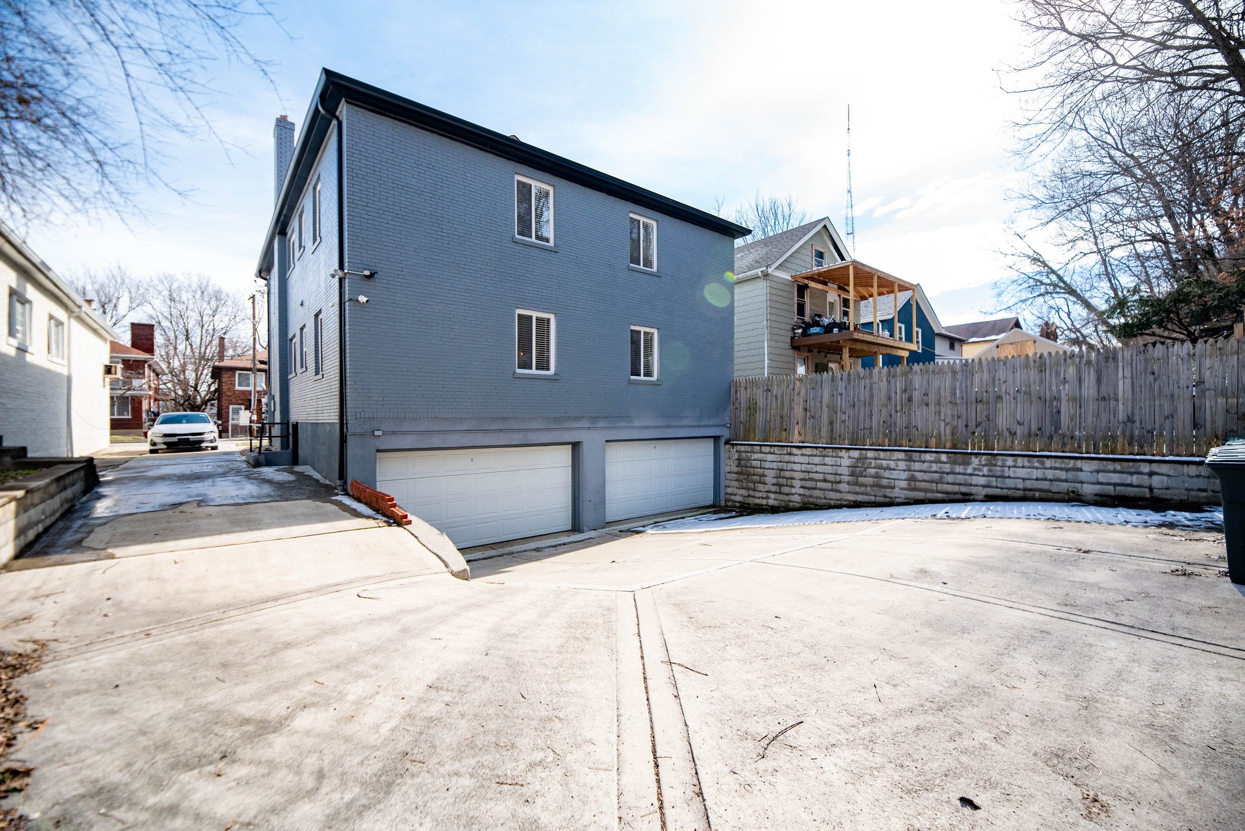 Side view of a gray two-story building with two garage doors, a sloped driveway, and a wooden fence, with other residential houses visible in the background.