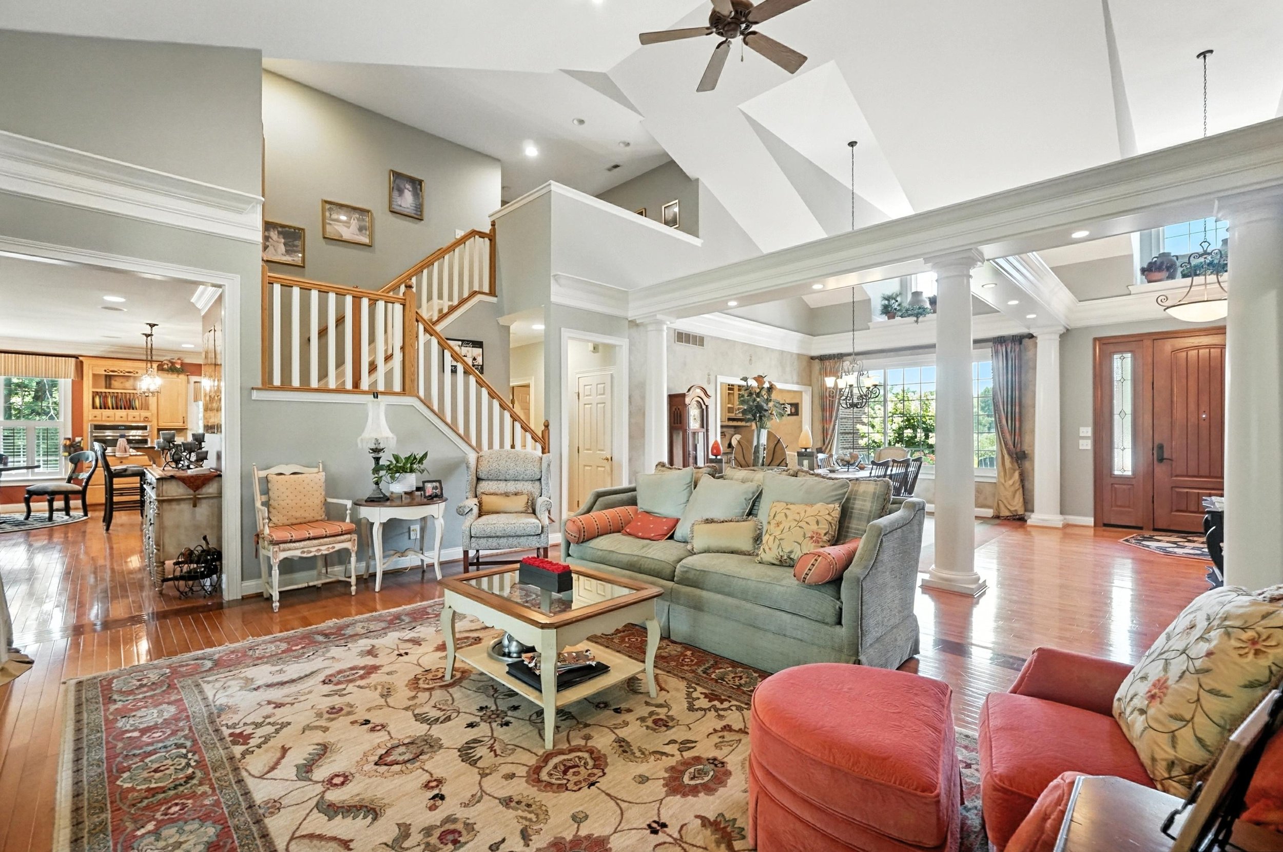 Spacious living room with hardwood floors, comfortable sofas, and a large area rug featuring floral patterns. There are decorative throw pillows on the couches and chairs, and a glass-top coffee table in the center. The room has a high ceiling with c