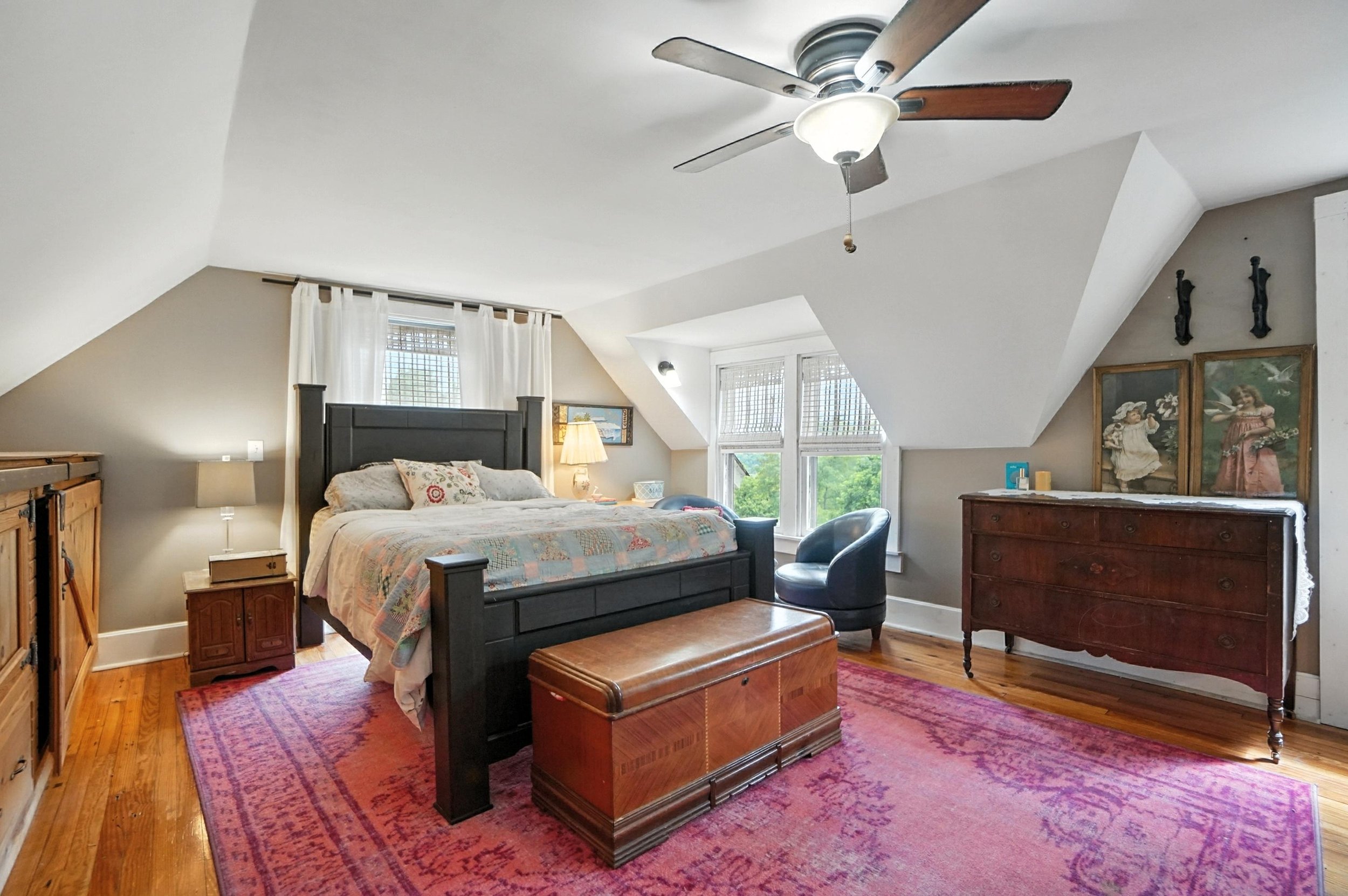 A cozy bedroom with a dark wooden bed frame, quilted bedspread, and decorative pillows. There is a pink rug on the wooden floor, a brown wooden trunk at the foot of the bed, two small wooden dressers, a black armchair near the window, and a small nig