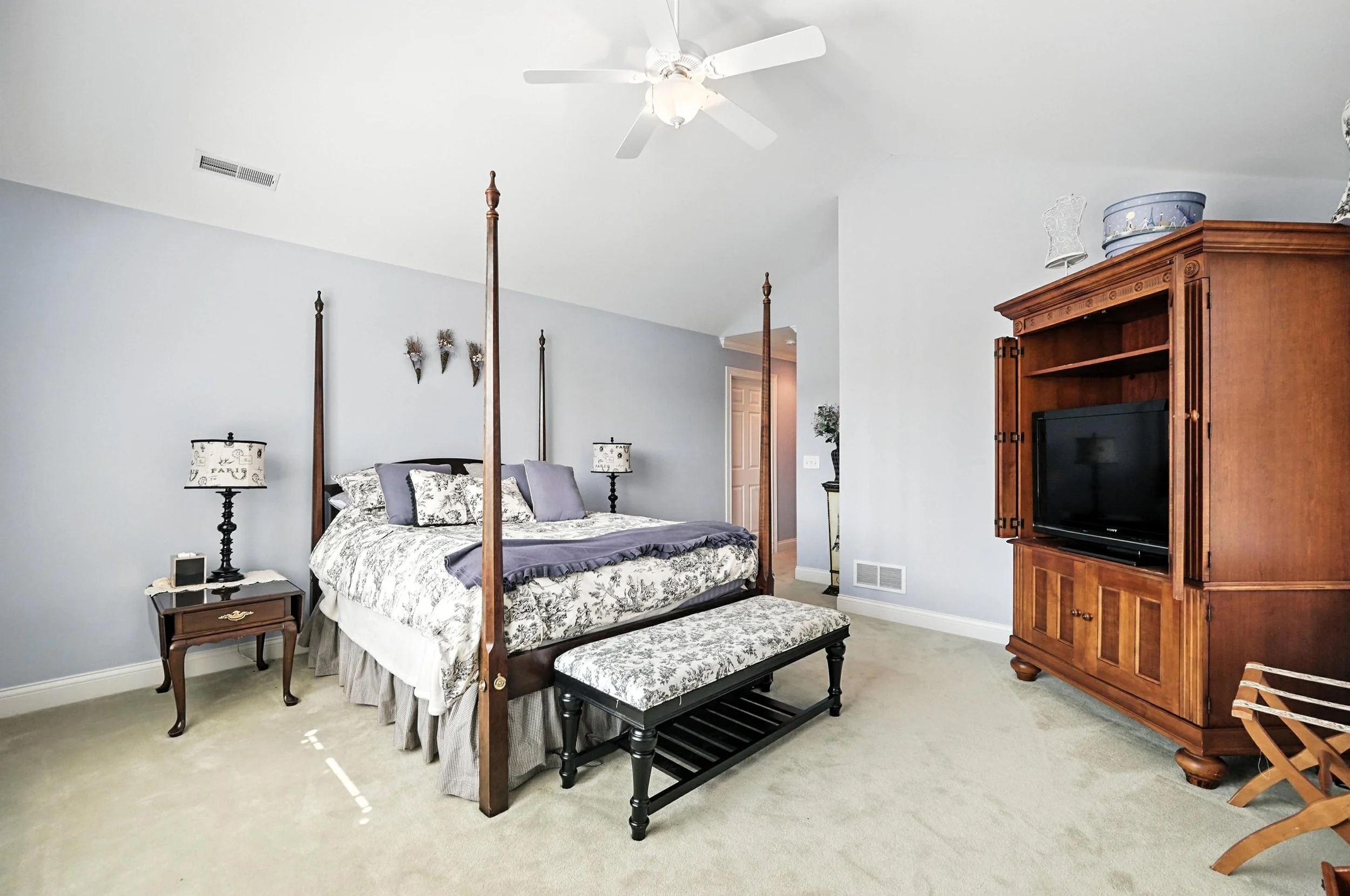 A bedroom with a four-poster bed, bedside tables with lamps, a patterned bench at the foot of the bed, a wooden entertainment center with a TV, and a ceiling fan.