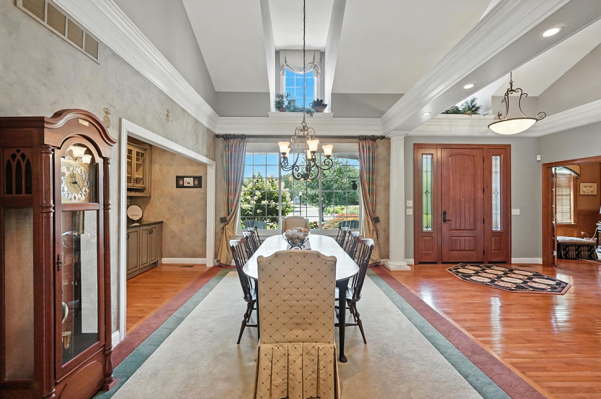 Dining room with wooden dining table, upholstered chairs, large window with curtains, chandelier, and a grandfather clock