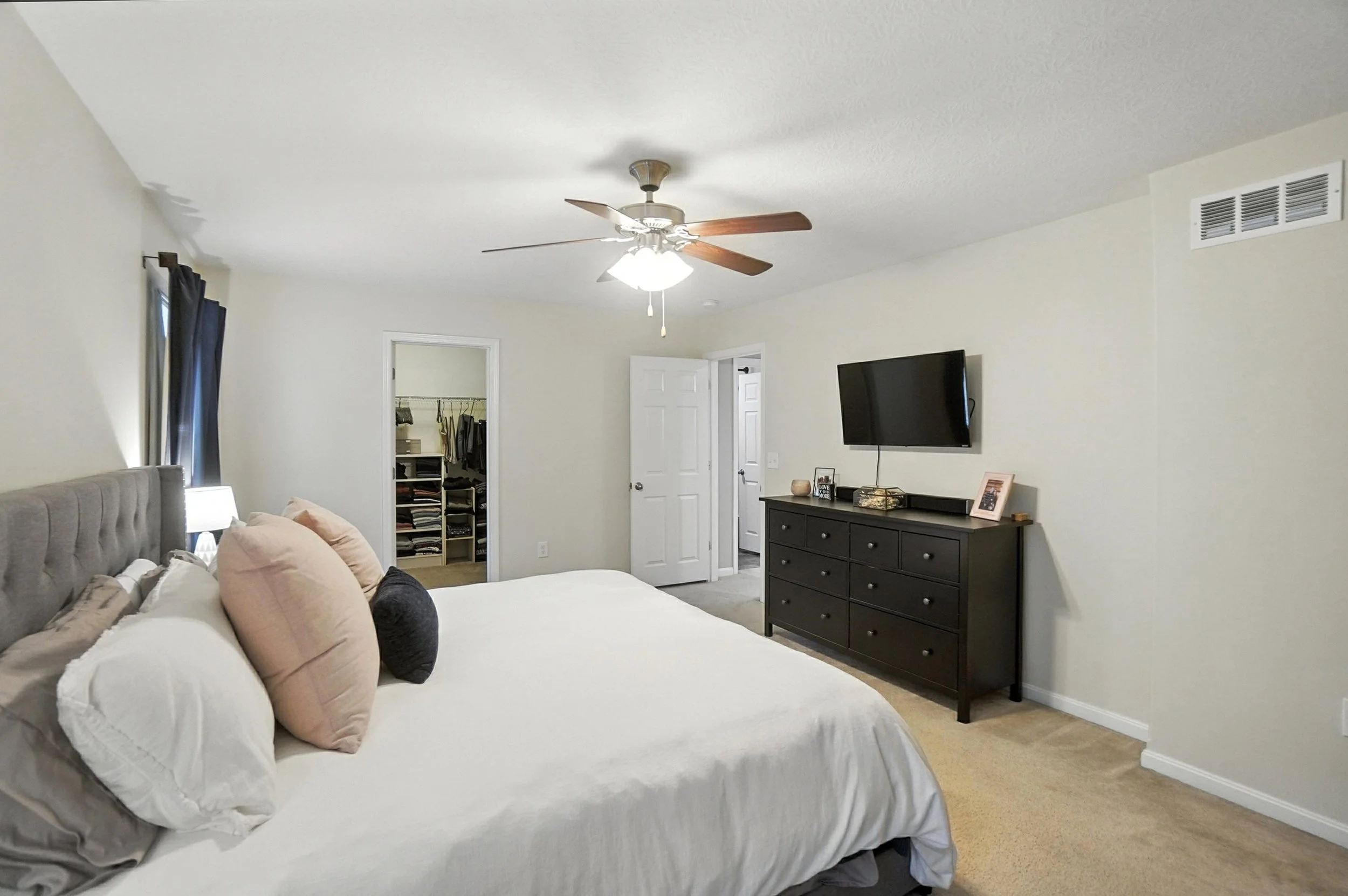 Bedroom with bed covered in white linens, multiple pillows, a dresser with a TV mounted above it, an open closet door, and a ceiling fan.