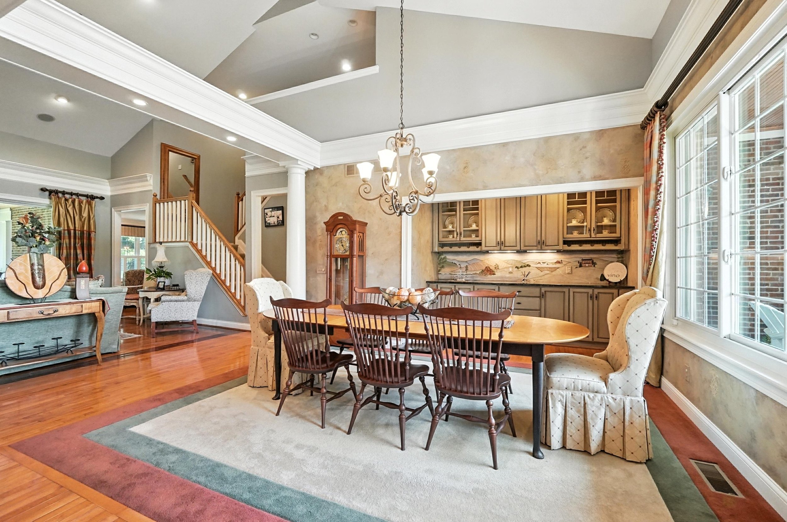 Dining room with wooden table, seven chairs, beige upholstered armchair, large window with patterned curtains, wooden hutch with dishes, chandelier, and hardwood floors.