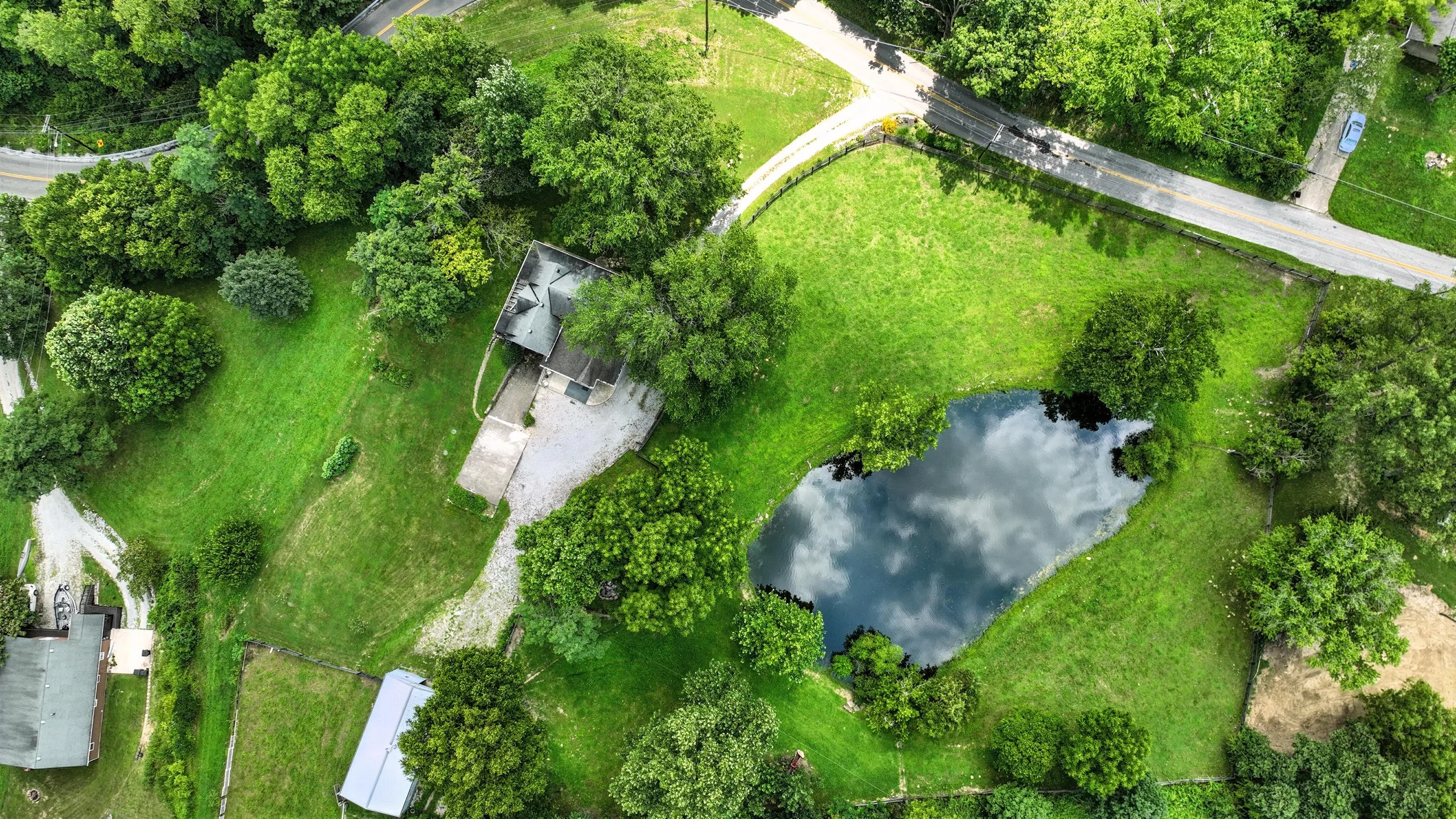 Aerial view of a backyard with a pond, lawn, trees, a house, and a gravel driveway.
