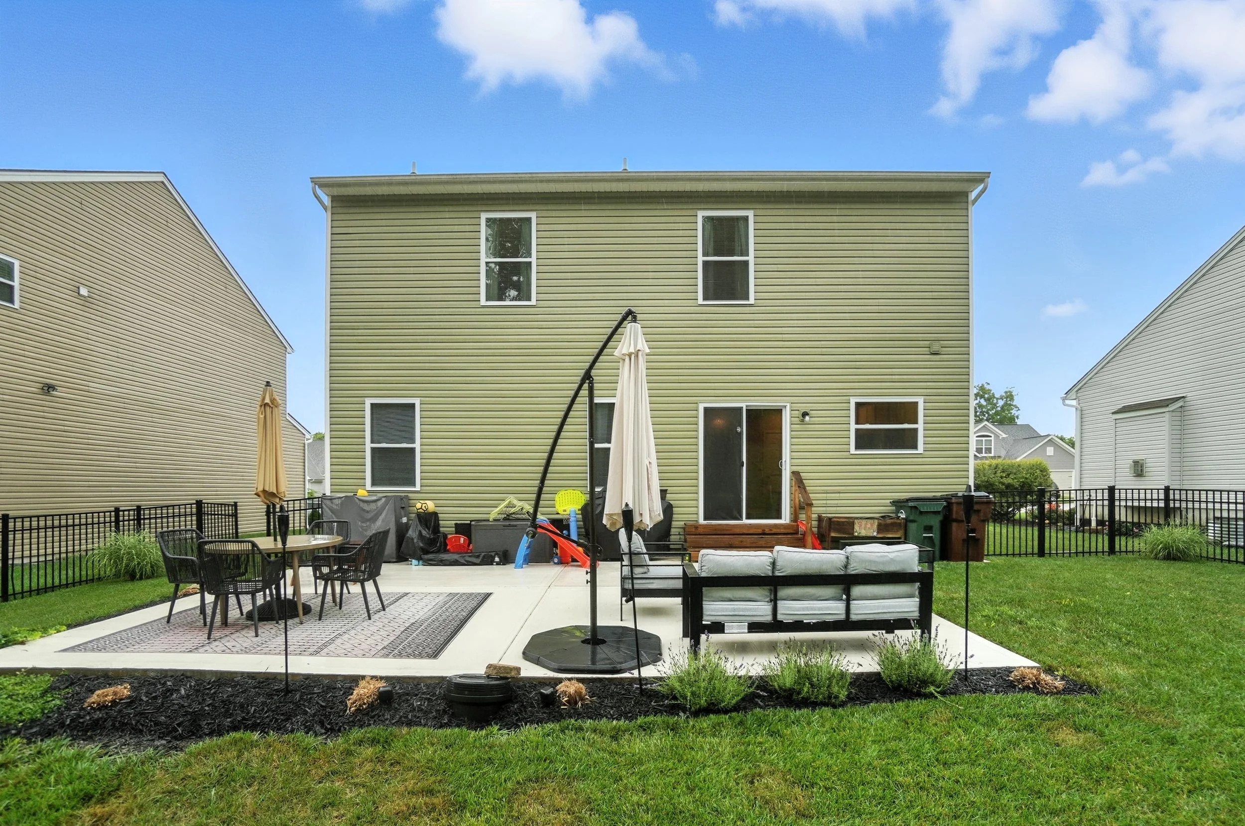 Backyard patio with outdoor furniture, a black metal fence, a house with yellow siding, and a blue sky with clouds.