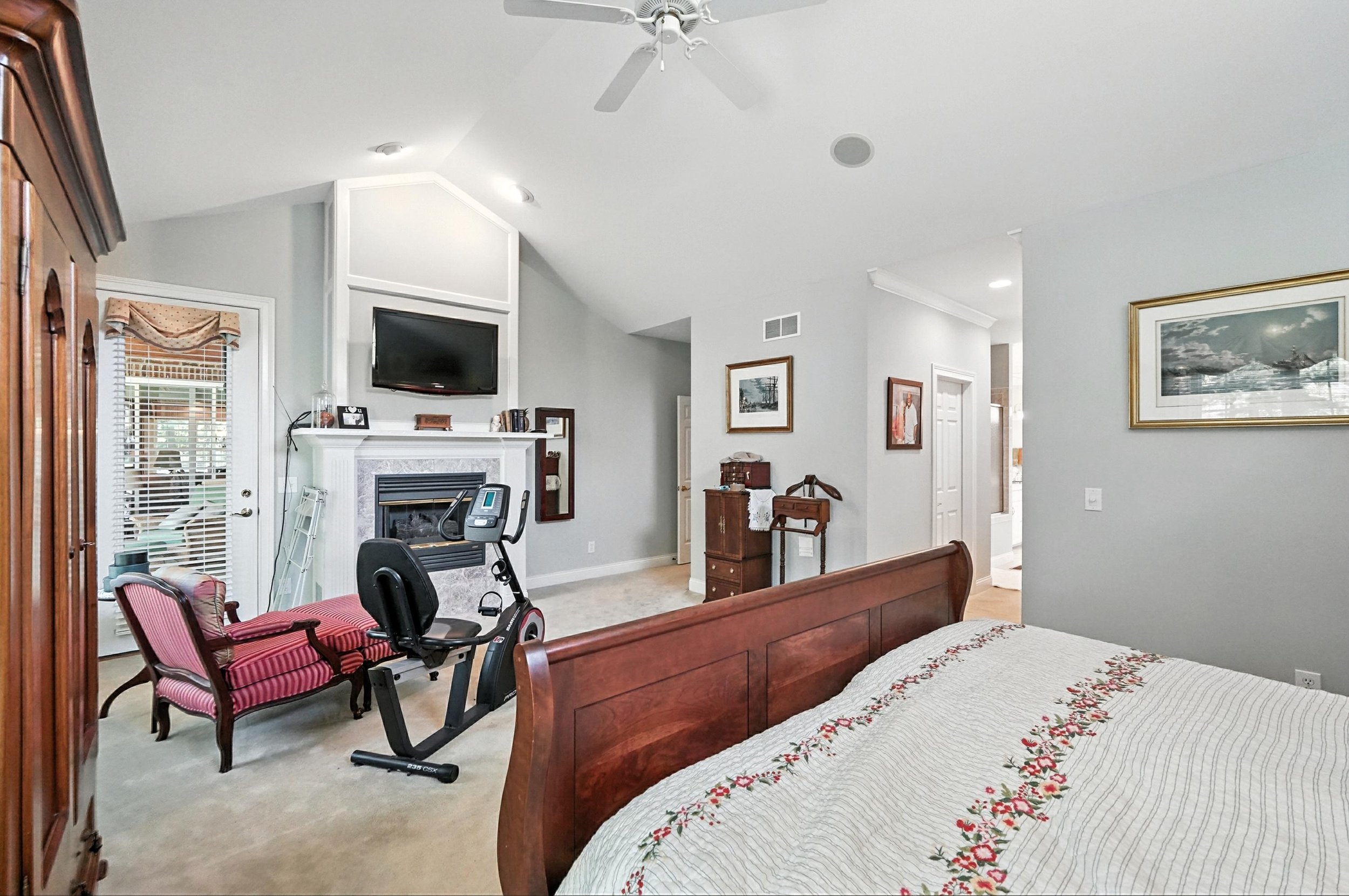 Interior of a bedroom with a wooden bed, quilted bedspread, exercise bike, upholstered chair, fireplace with a TV above it, and various artwork on the walls.
