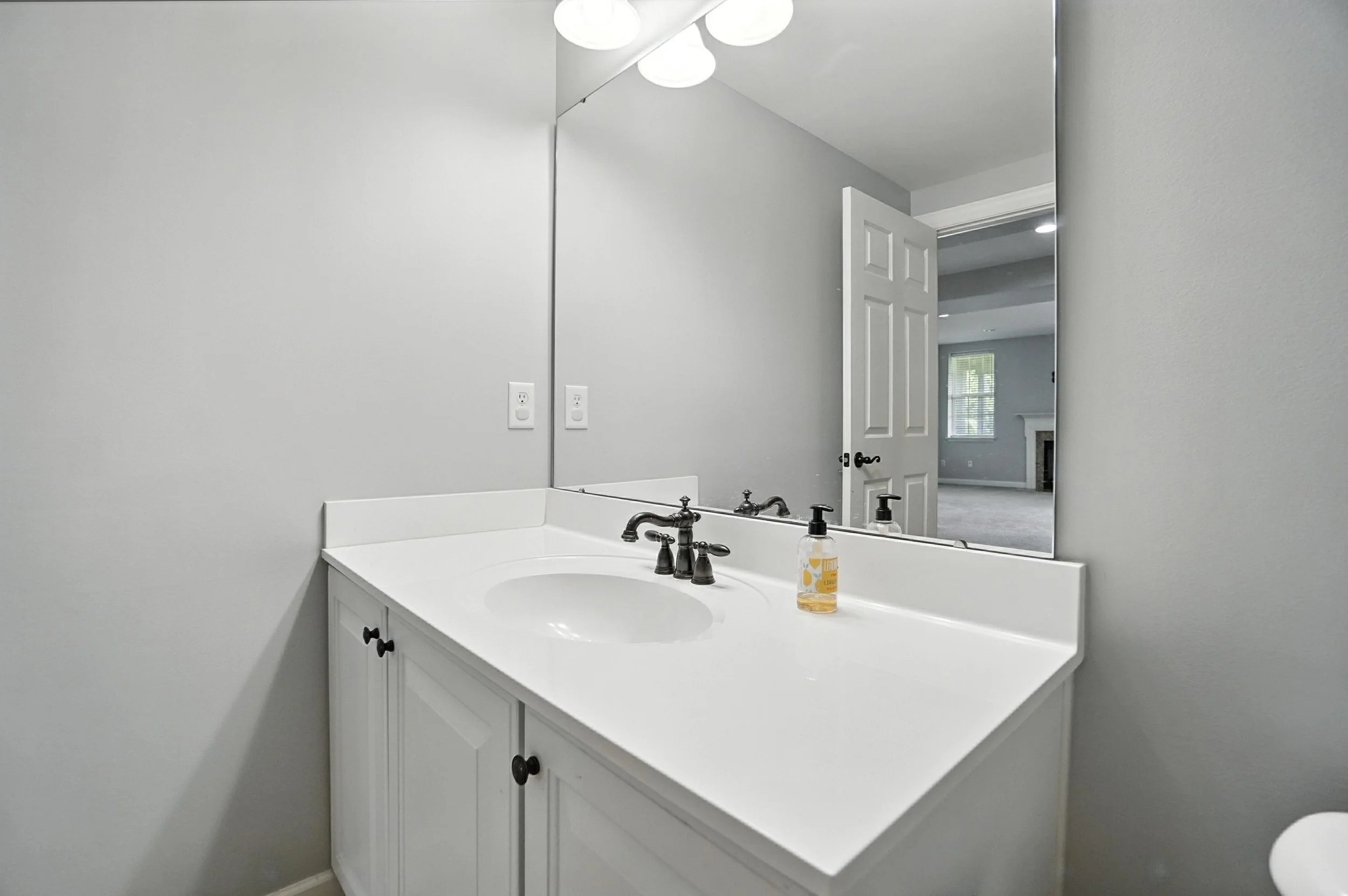 Bathroom vanity with white countertop, a large mirror, soap dispenser, and a partially open door leading to a living room with a fireplace and window.