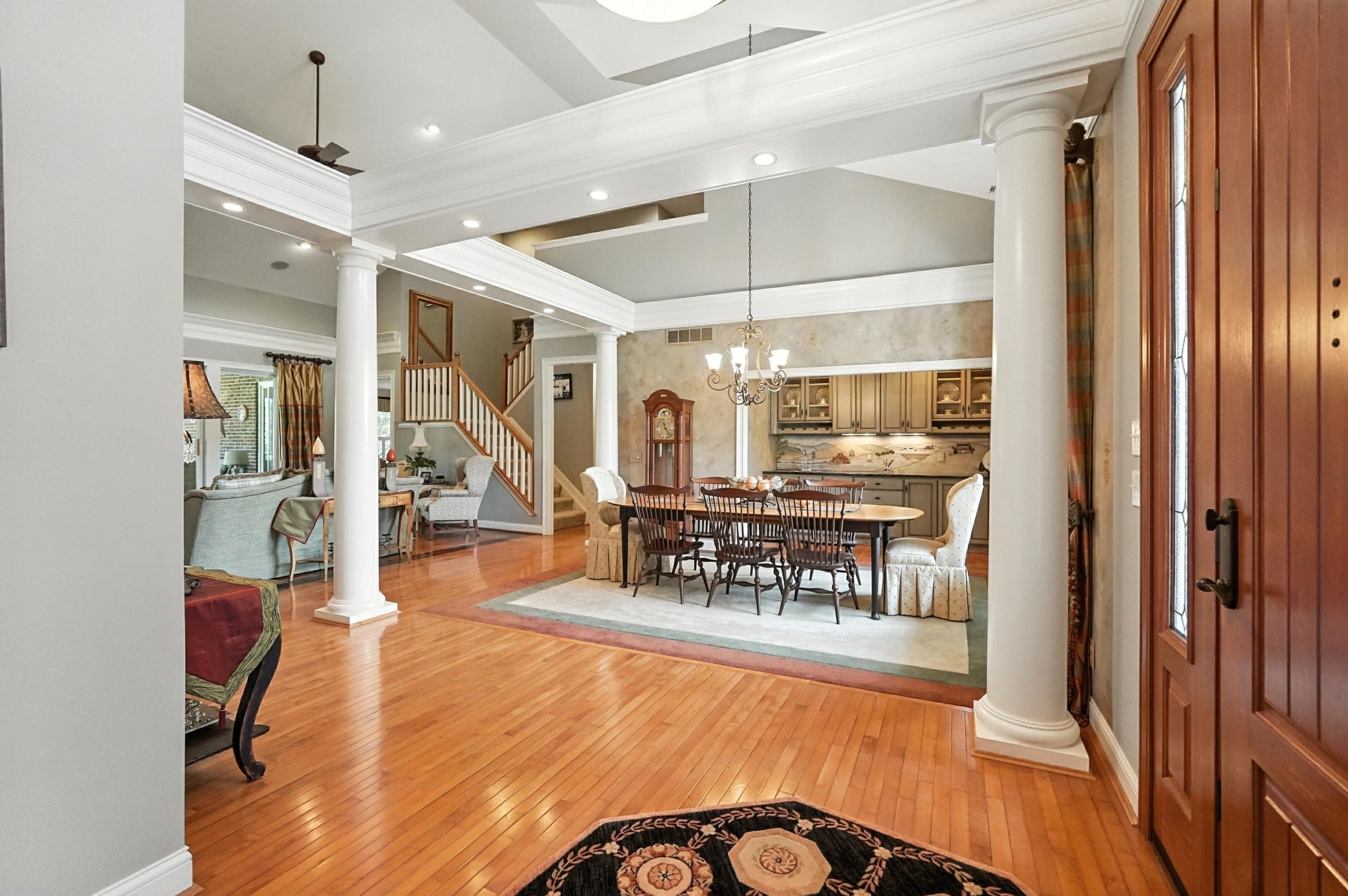 View of a spacious dining area and living room in a home with hardwood floors, white columns, a chandelier, and a staircase.