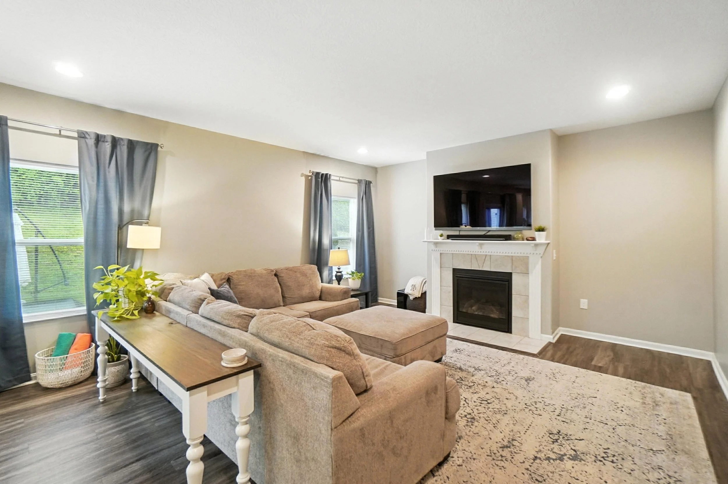 Living room with beige sectional sofa, patterned area rug, fireplace with mounted TV, two windows with blue curtains, and various potted plants.