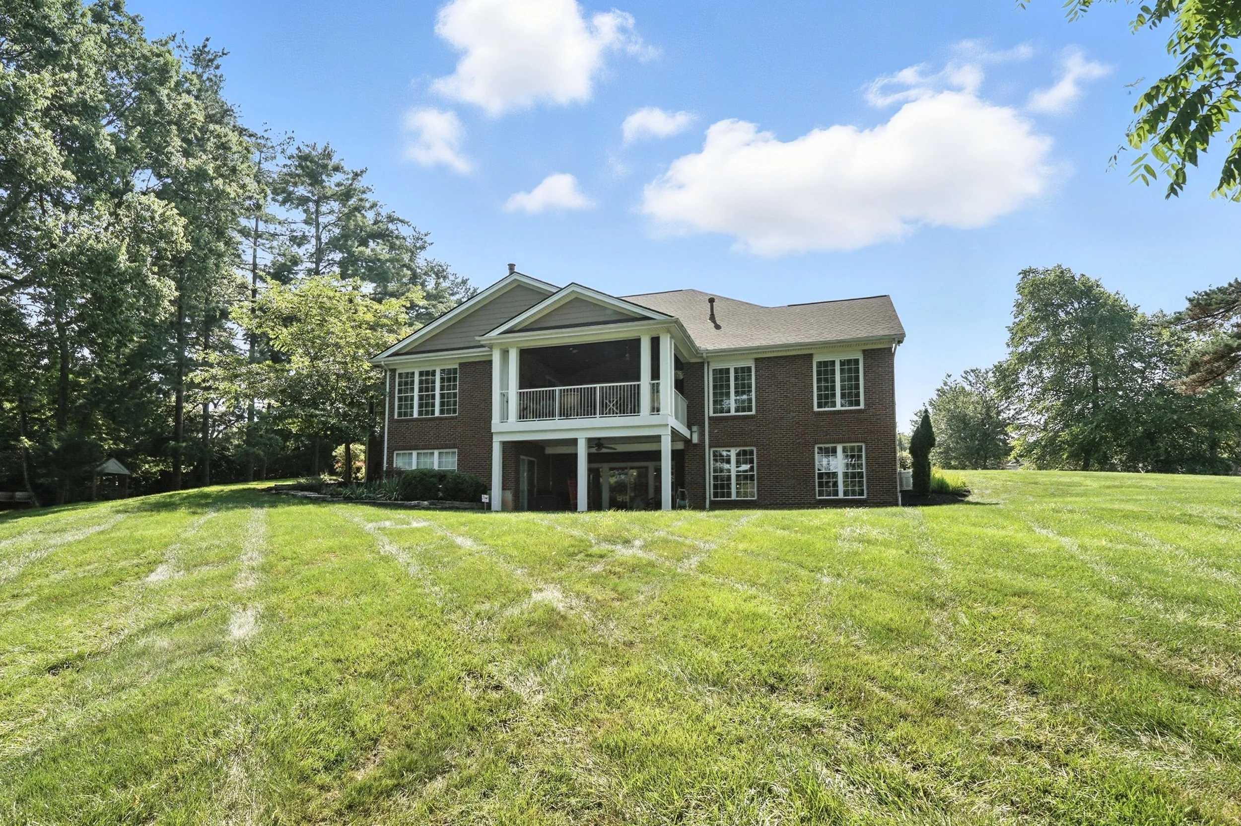 Large brick house with a screened porch and balcony, surrounded by a green lawn and trees under a partly cloudy sky.