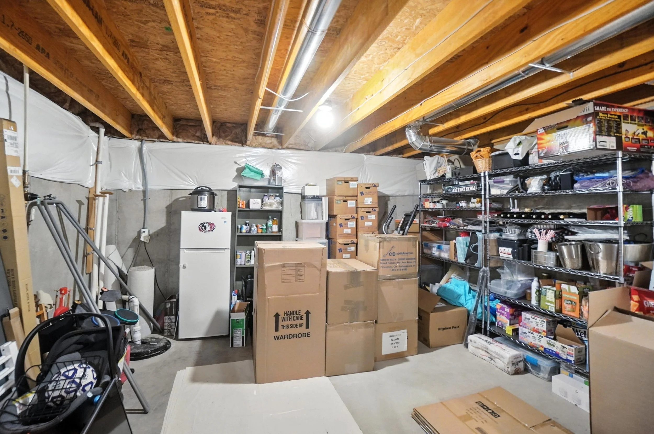 A cluttered basement storage room with cardboard boxes, metal shelving filled with various household items, a small refrigerator, and a laundry basket.