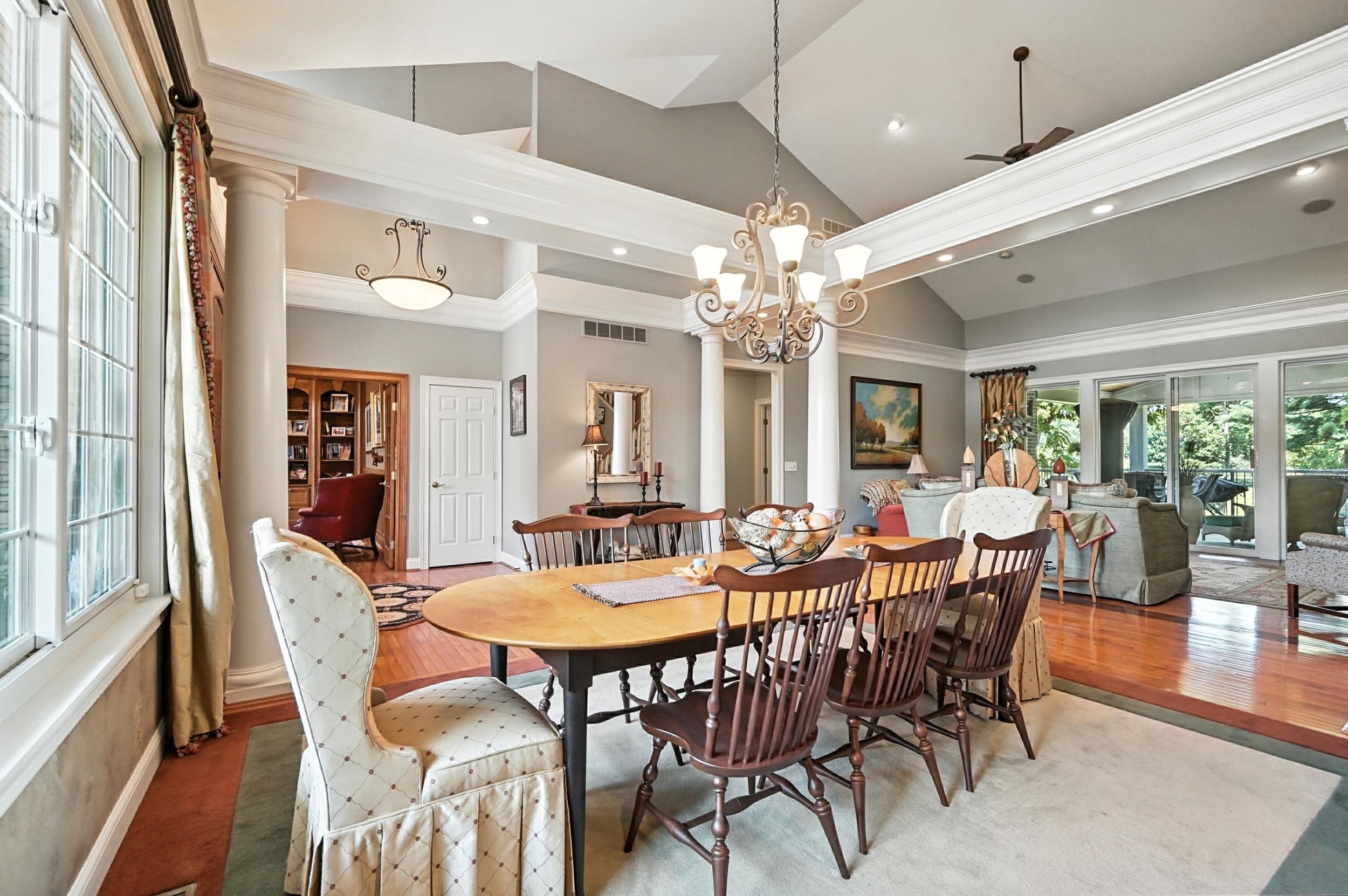Open-concept living and dining room with wooden flooring, light gray walls, large windows, and elegant furniture including a dining table with mix of chairs, sofas, and decorative elements.