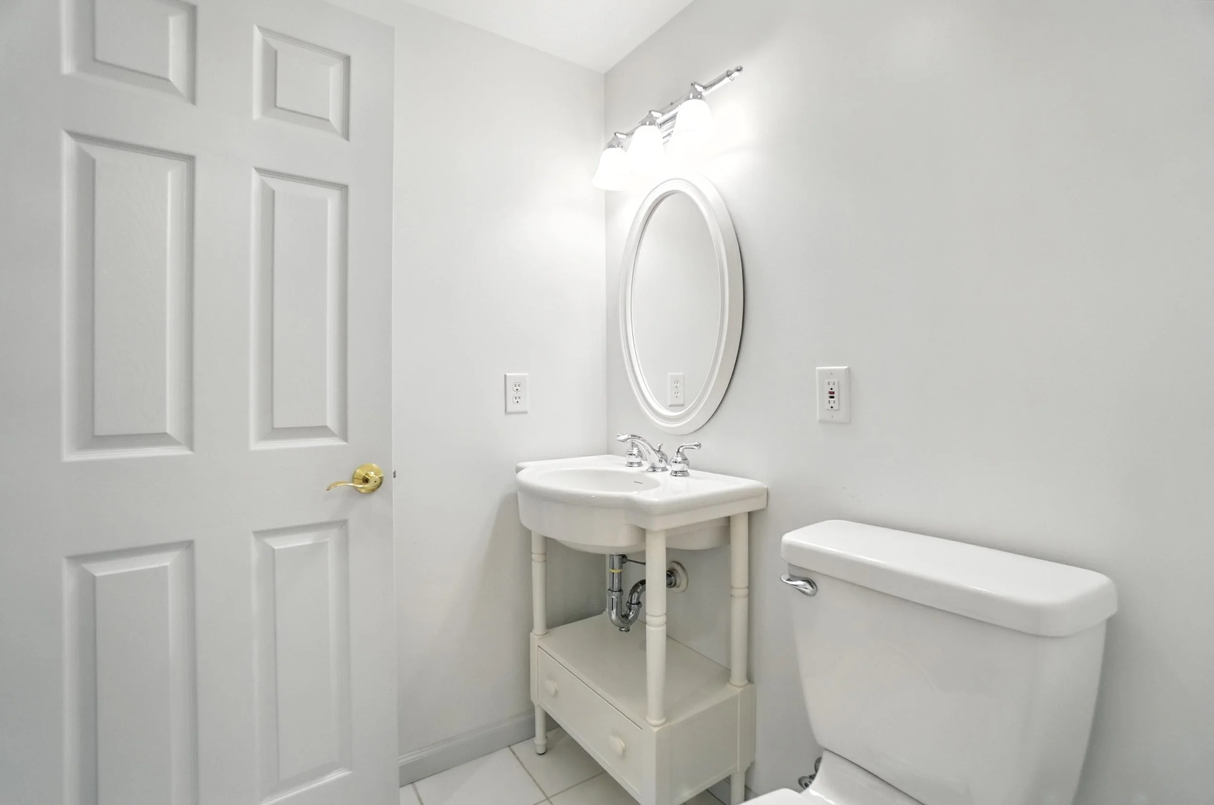 Small white bathroom with a door on the left, a pedestal sink with a mirror above, a light fixture, and a toilet on the right side.
