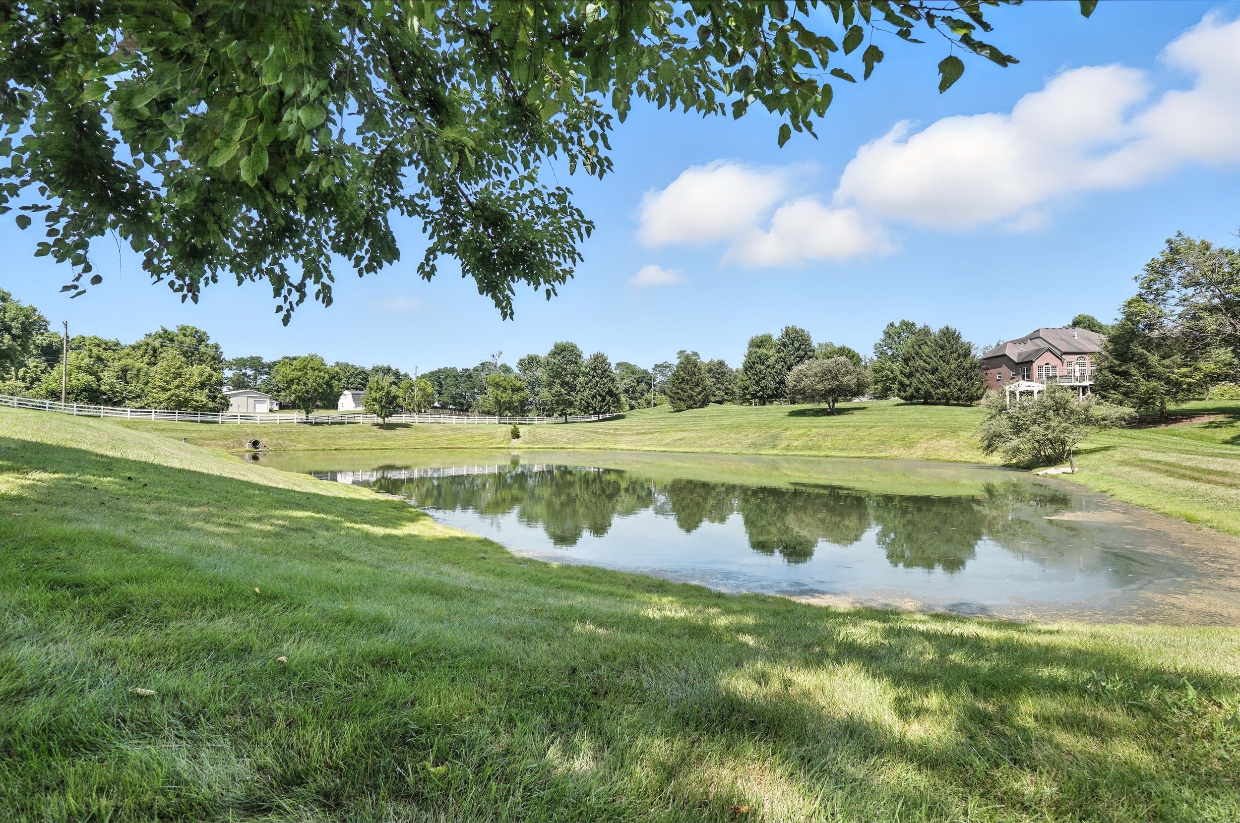 A peaceful landscape featuring a small pond reflecting the blue sky and white clouds, surrounded by green grass, trees, and a house on the right side in the distance.