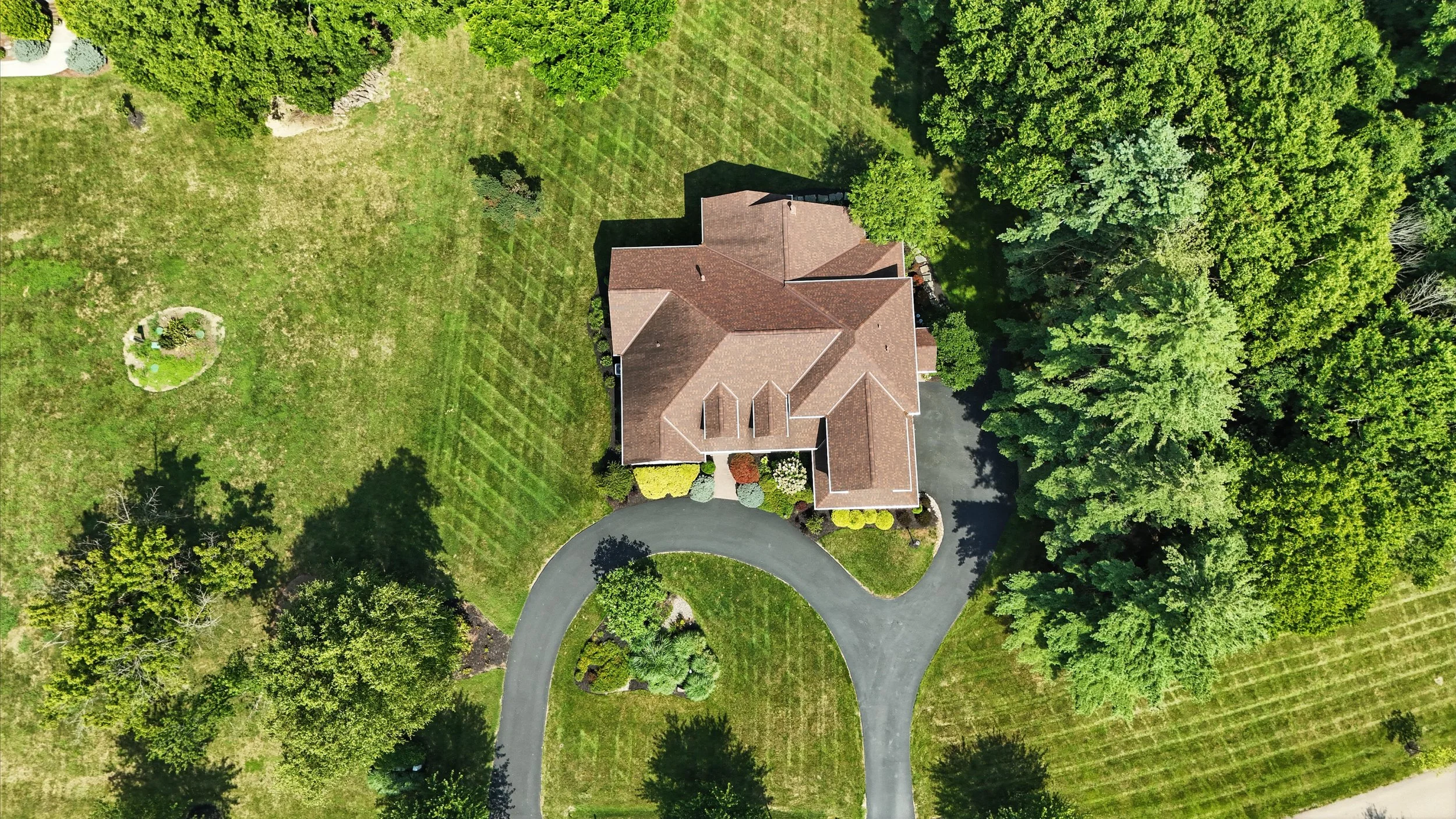 Aerial view of a large house with a brown roof, surrounded by a green lawn with trees and a circular driveway.