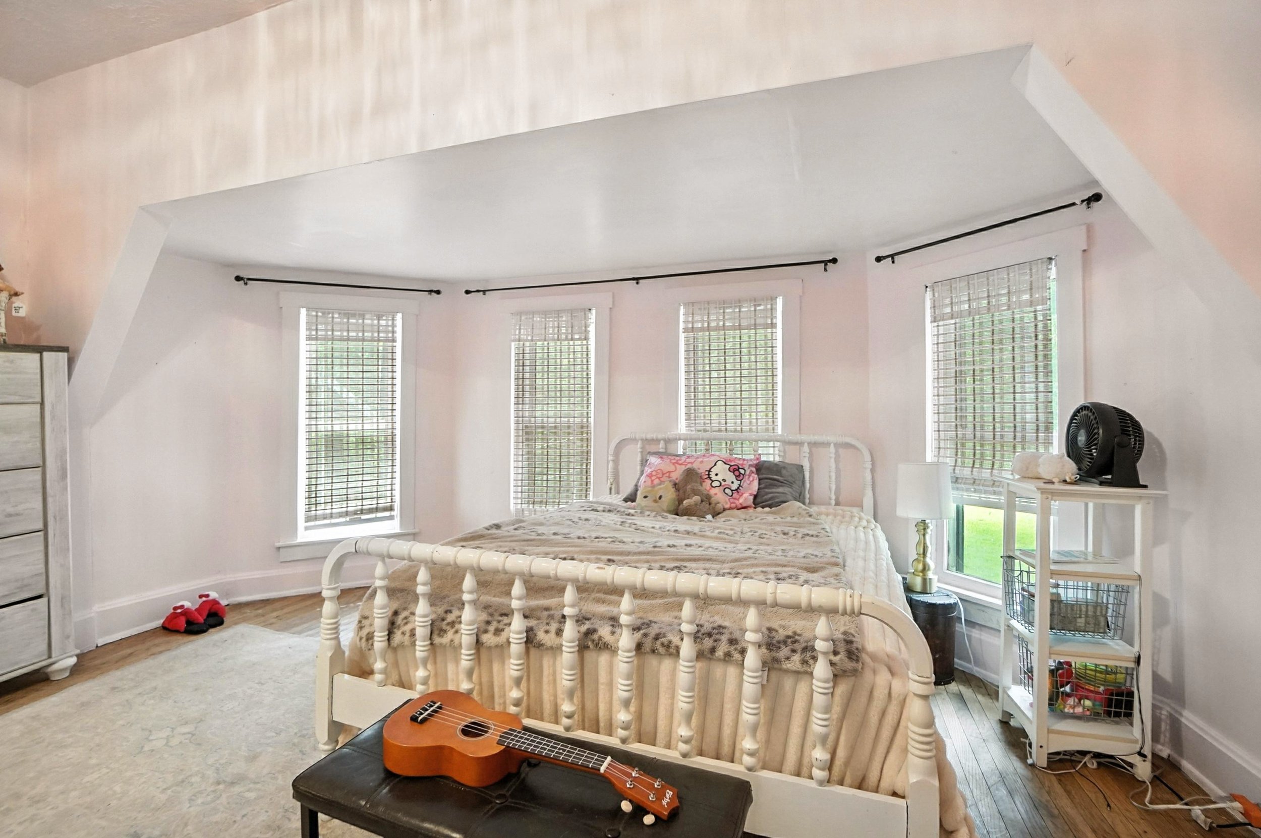 A bedroom with a white iron bed, multiple windows with blinds, a guitar on a small table, and a white shelf with a fan and items on it.