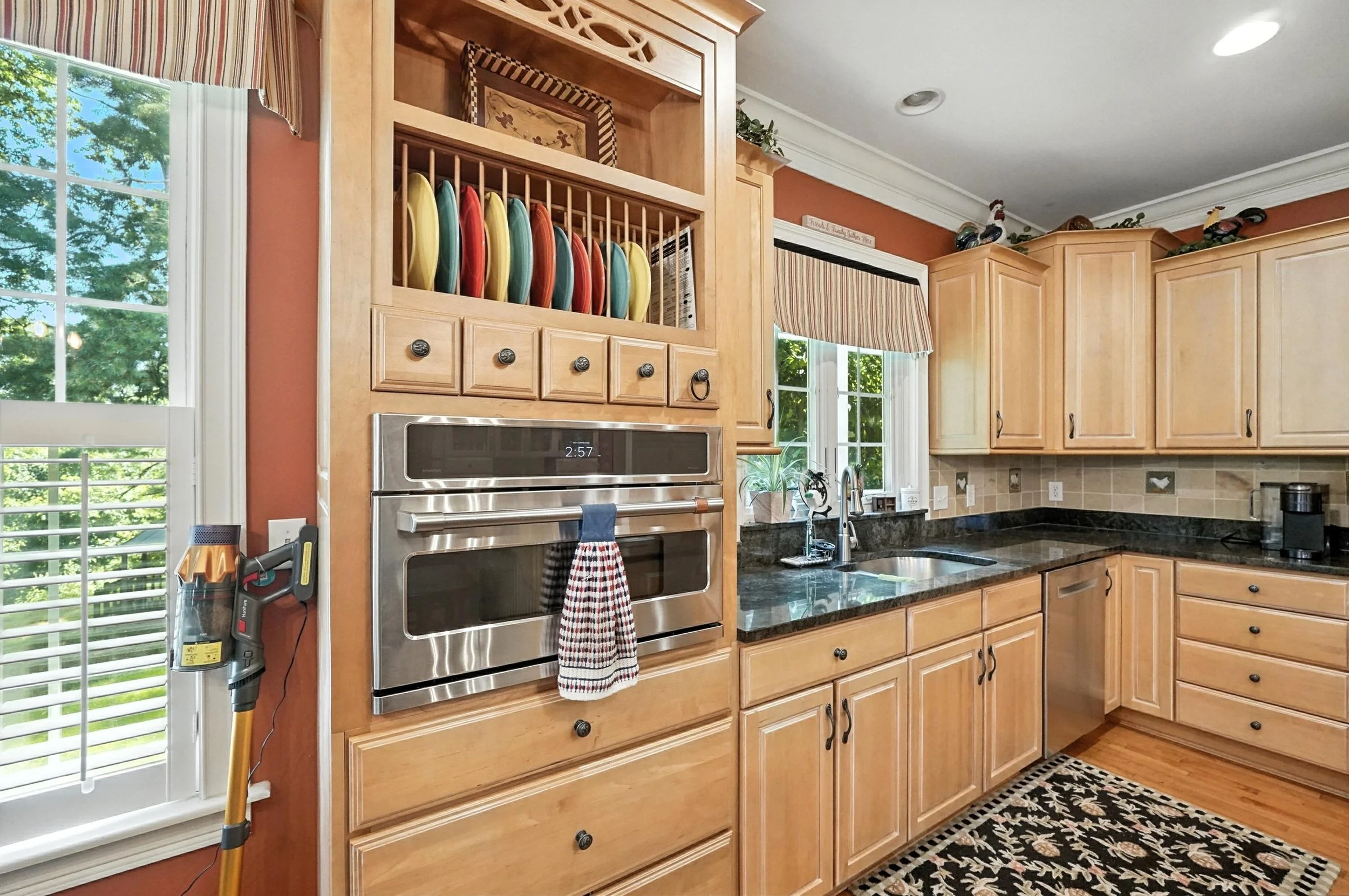 Kitchen with wooden cabinets, black granite countertops, stainless steel double oven, dish rack with colorful plates above oven, windows with striped valances, and a black patterned rug on a hardwood floor.