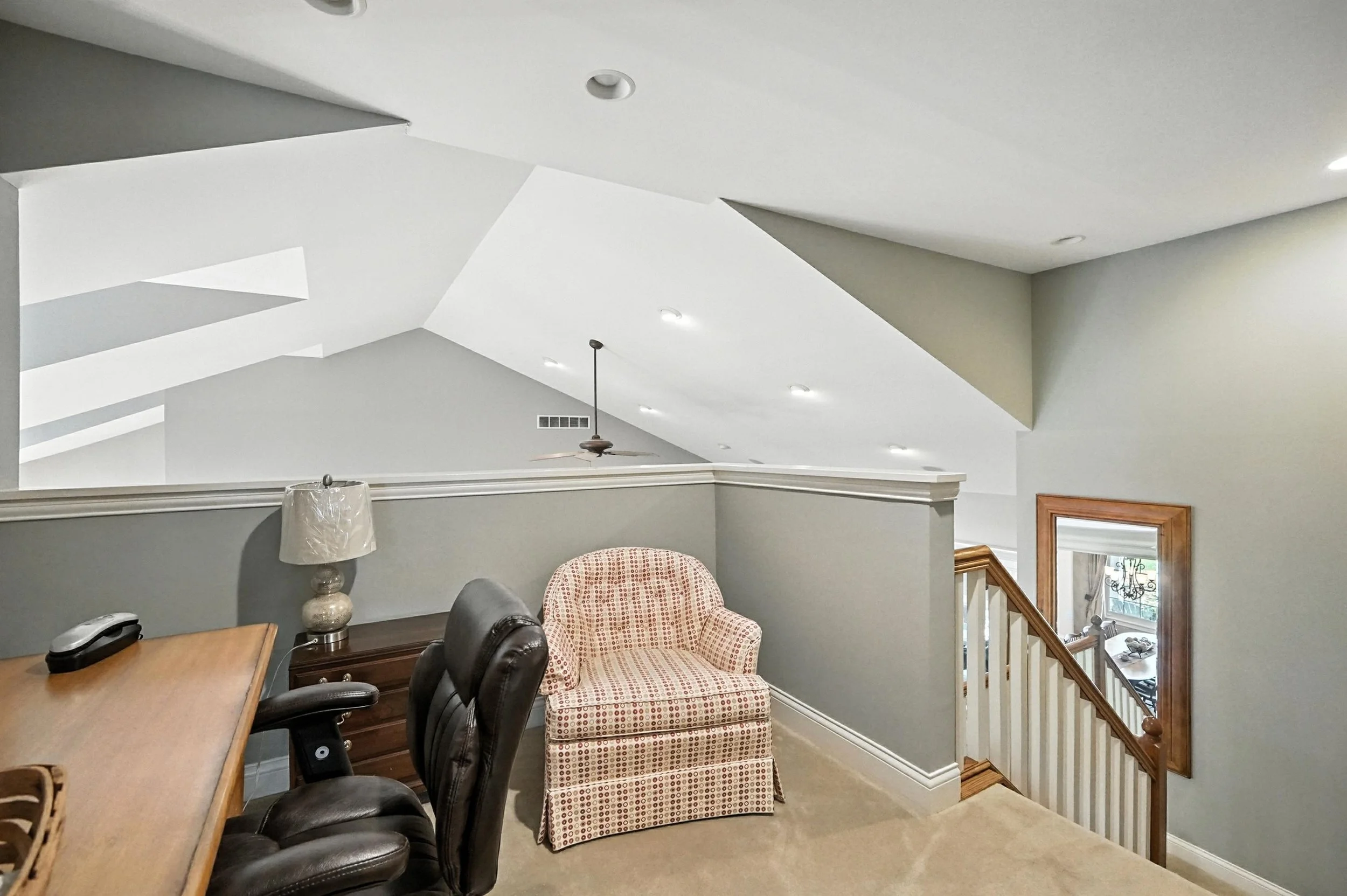 Interior view of a second-floor landing with office chair, side table, table lamp, patterned armchair, staircase with wooden handrail, and ceiling with multiple light fixtures and a ceiling fan.