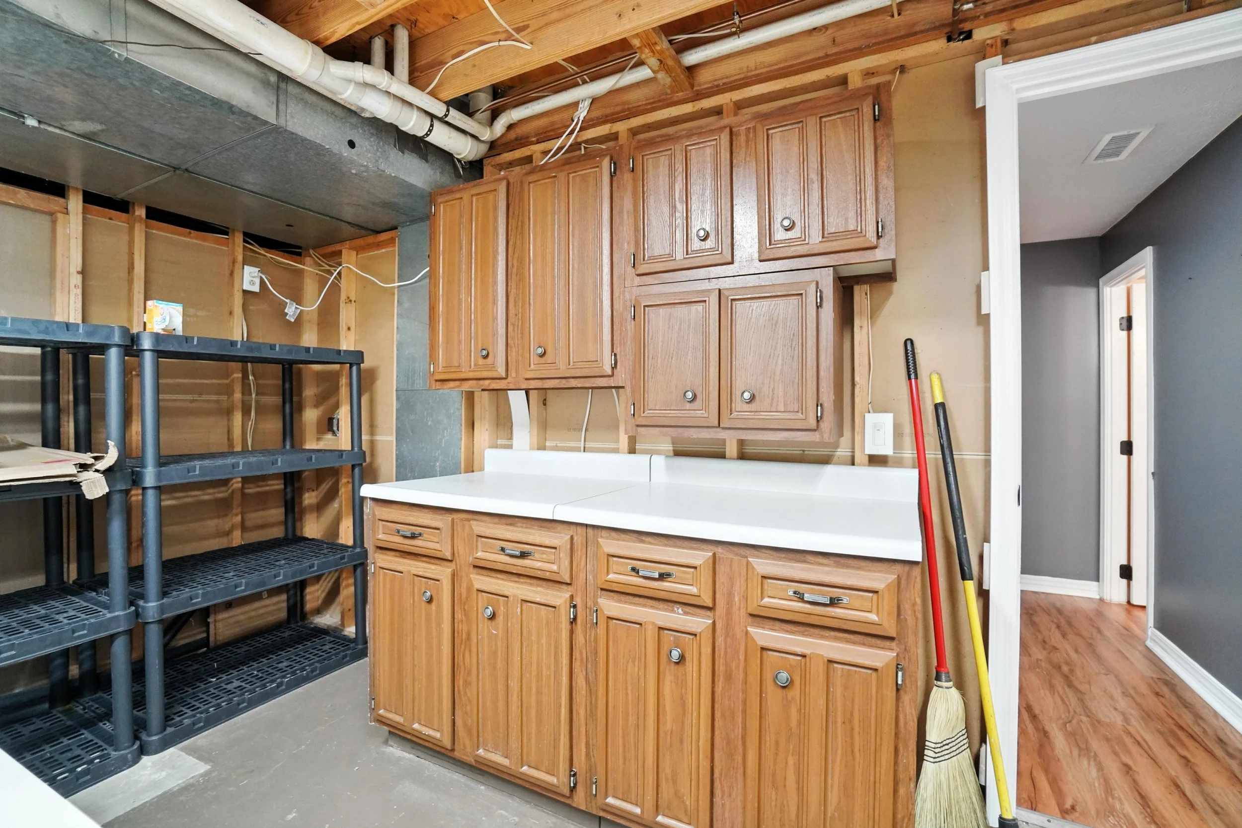 Kitchen with wooden cabinets, a white countertop, cleaning tools, and an open wall showing exposed pipes and wooden framing.