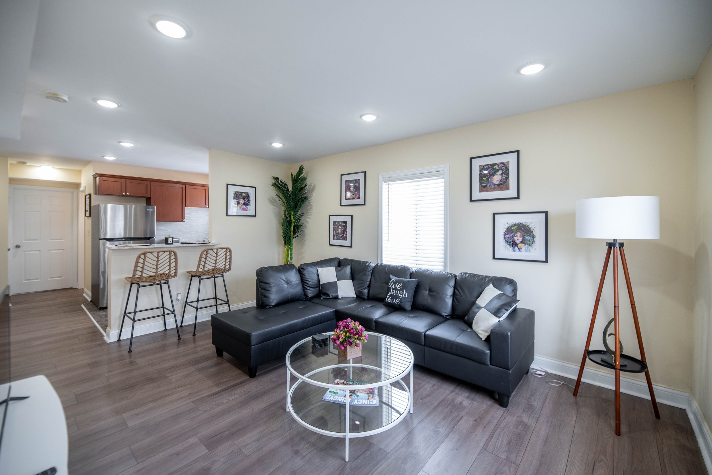 Modern living room with black leather sectional sofa, circular glass coffee table, wall art, floor lamp, and open view to the kitchen with bar stools.