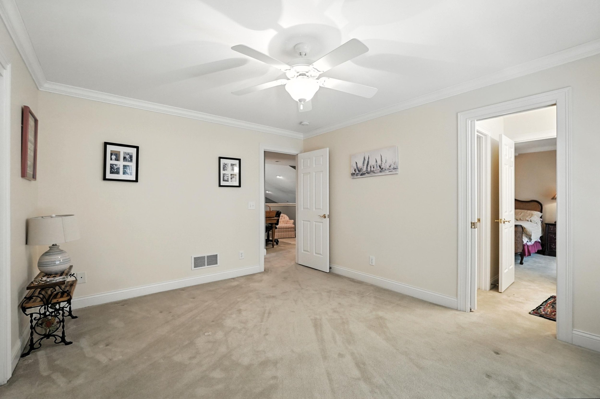 Empty bedroom with beige carpet, off-white walls, a ceiling fan with light, and two open doorways leading to other rooms. A small table with a lamp and a small picture frame is in the corner.