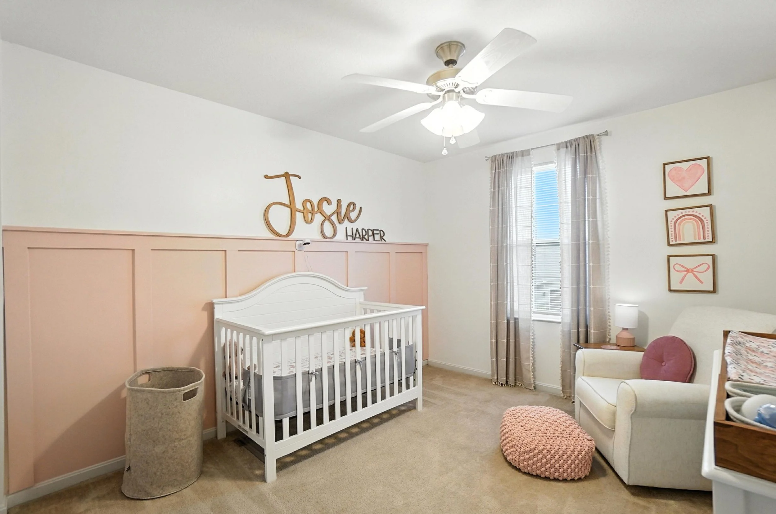 Nursery room with pink and white decor, a white crib, a beige armchair with a pink pillow, wall art with pinks and rainbows, and a window with blinds and tan curtains.