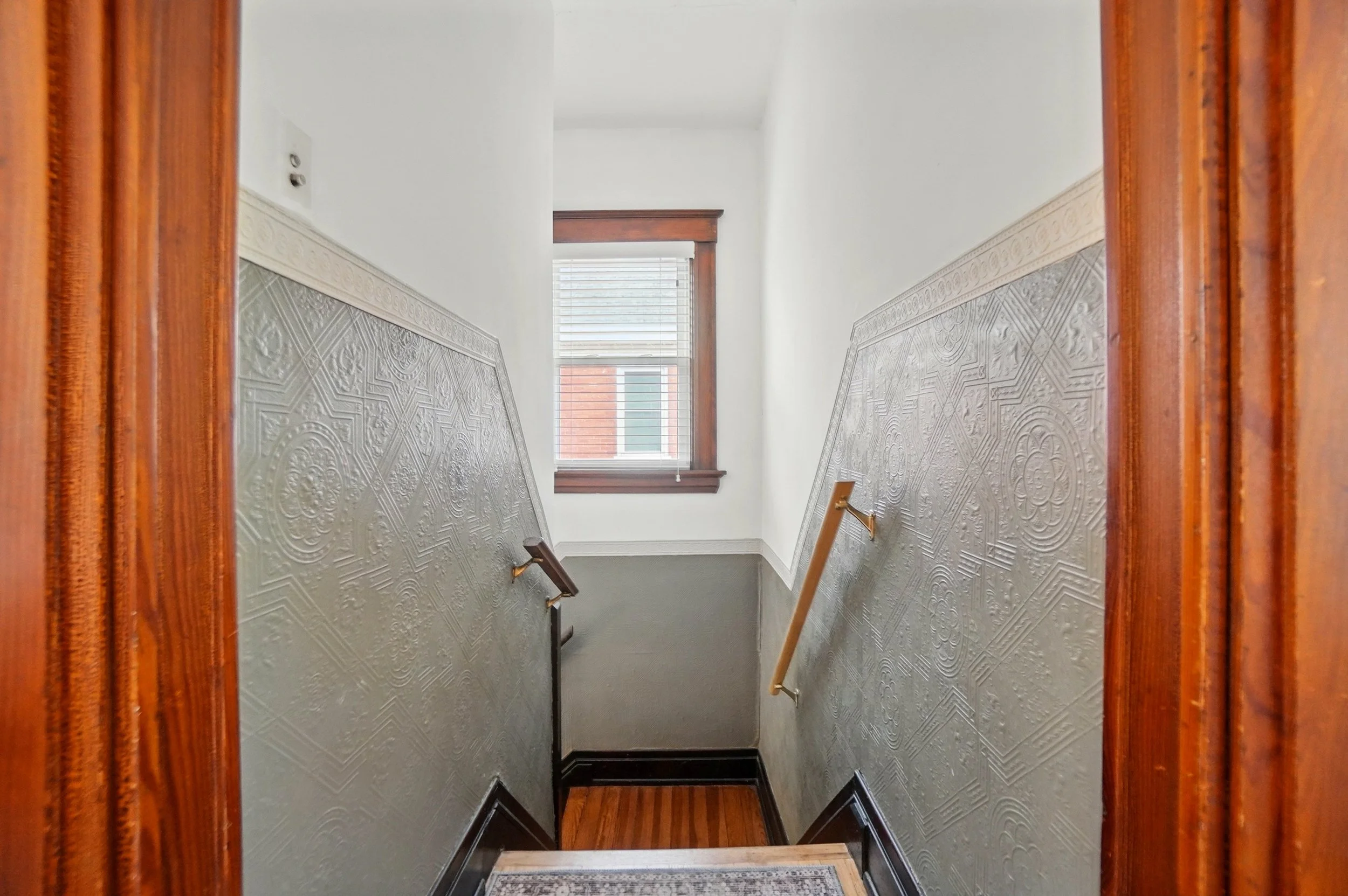 View of a staircase from the top, showing a window at the bottom, wood trim, and steel wainscoting with a decorative pattern.