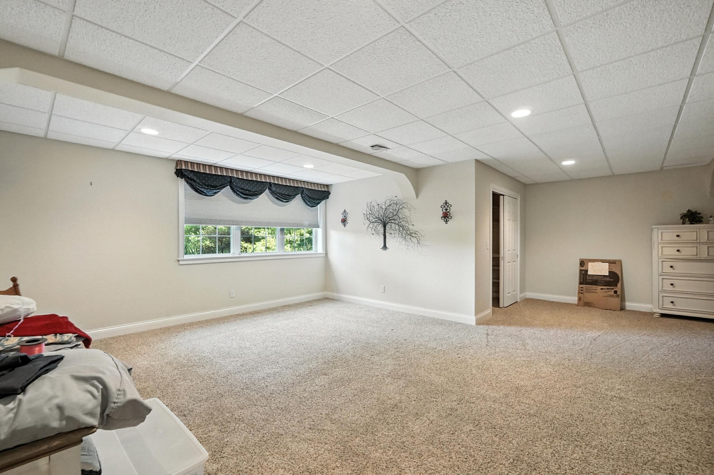 Empty basement room with beige carpet, white walls, a window with black and beige valance, wall decorations, and a white dresser.
