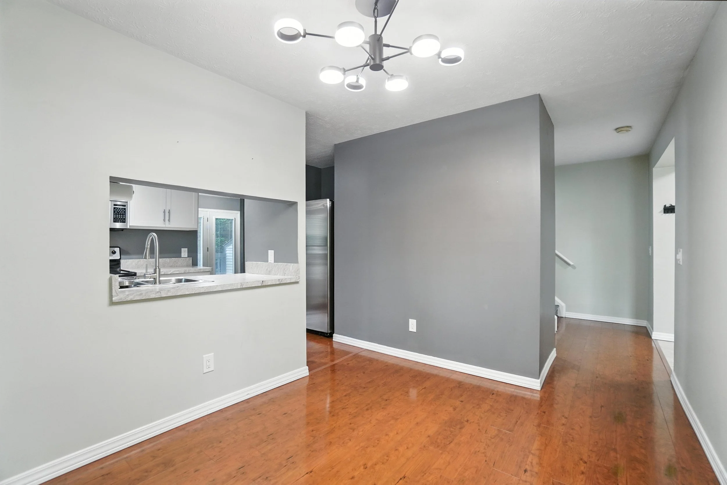Empty living room with gray and beige walls, hardwood floors, a ceiling light fixture, and an open kitchen with a pass-through window.