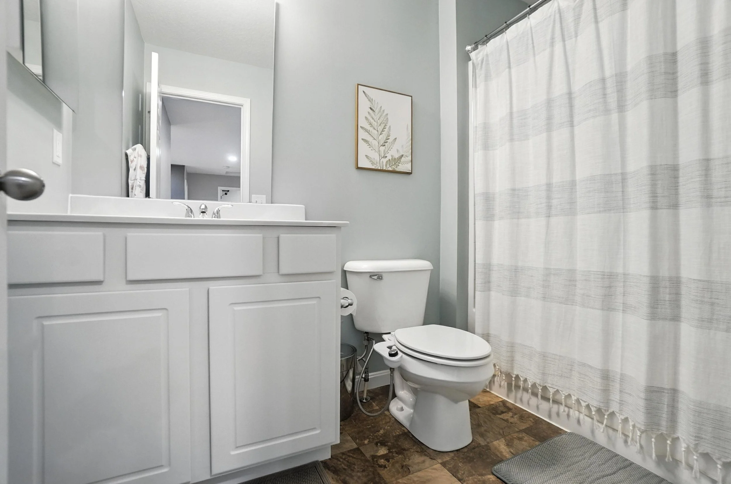 A clean bathroom with a white vanity, a toilet, a framed botanical print on the wall, and a shower with a striped curtain.