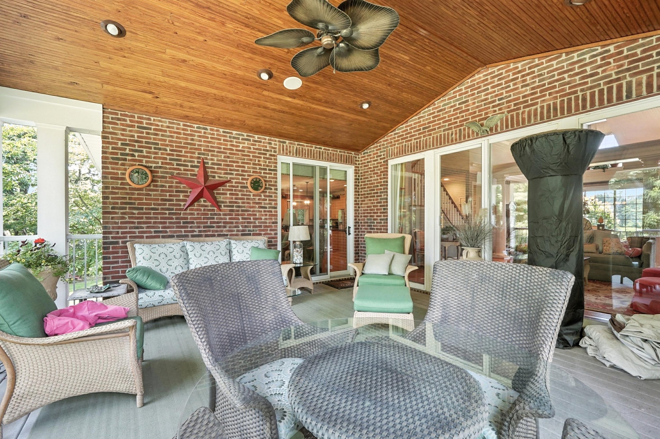 Covered outdoor porch with wicker and cushioned seating, brick wall, wood-paneled ceiling, ceiling fan, and glass sliding doors leading to indoor living space.