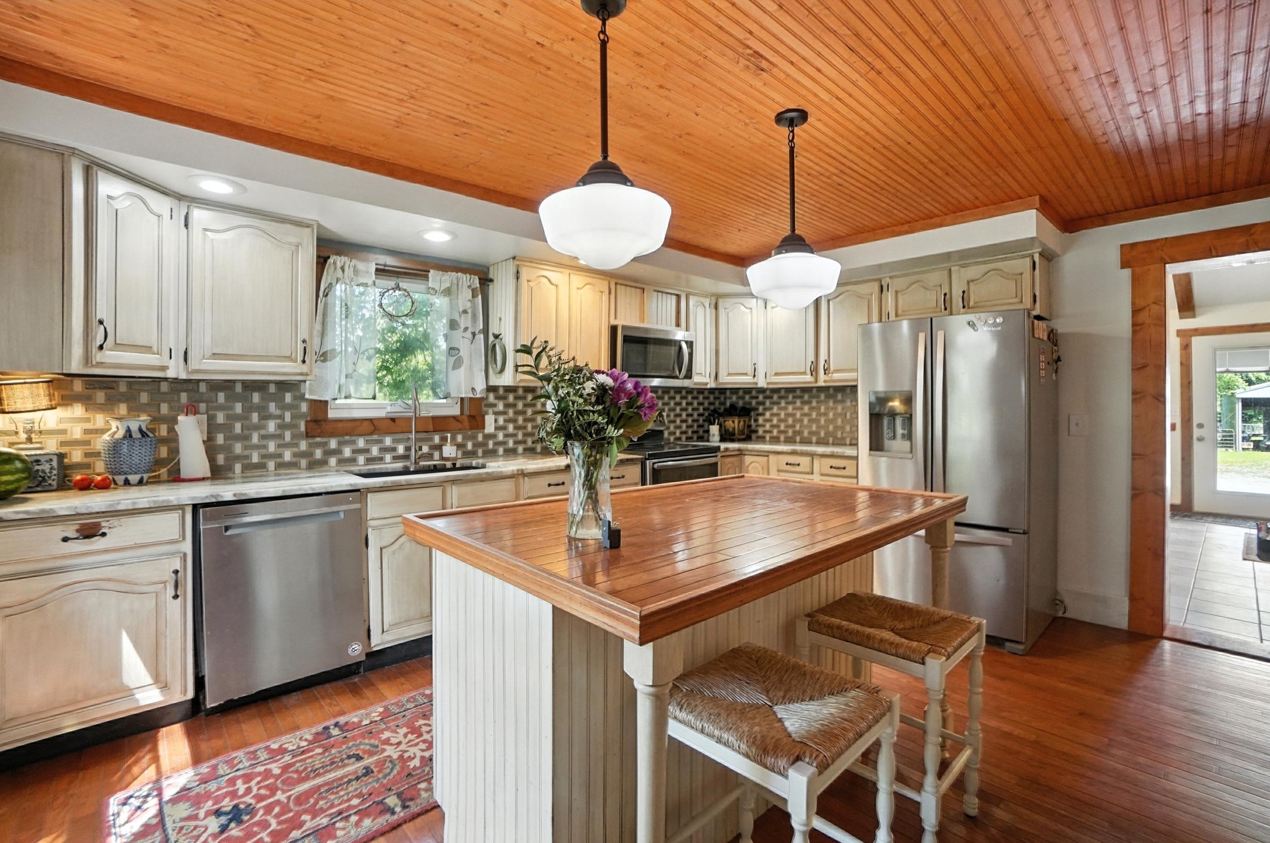 Cozy kitchen with white cabinets, a wooden ceiling, a kitchen island with a vase of flowers, stainless steel appliances, and a window with curtains.