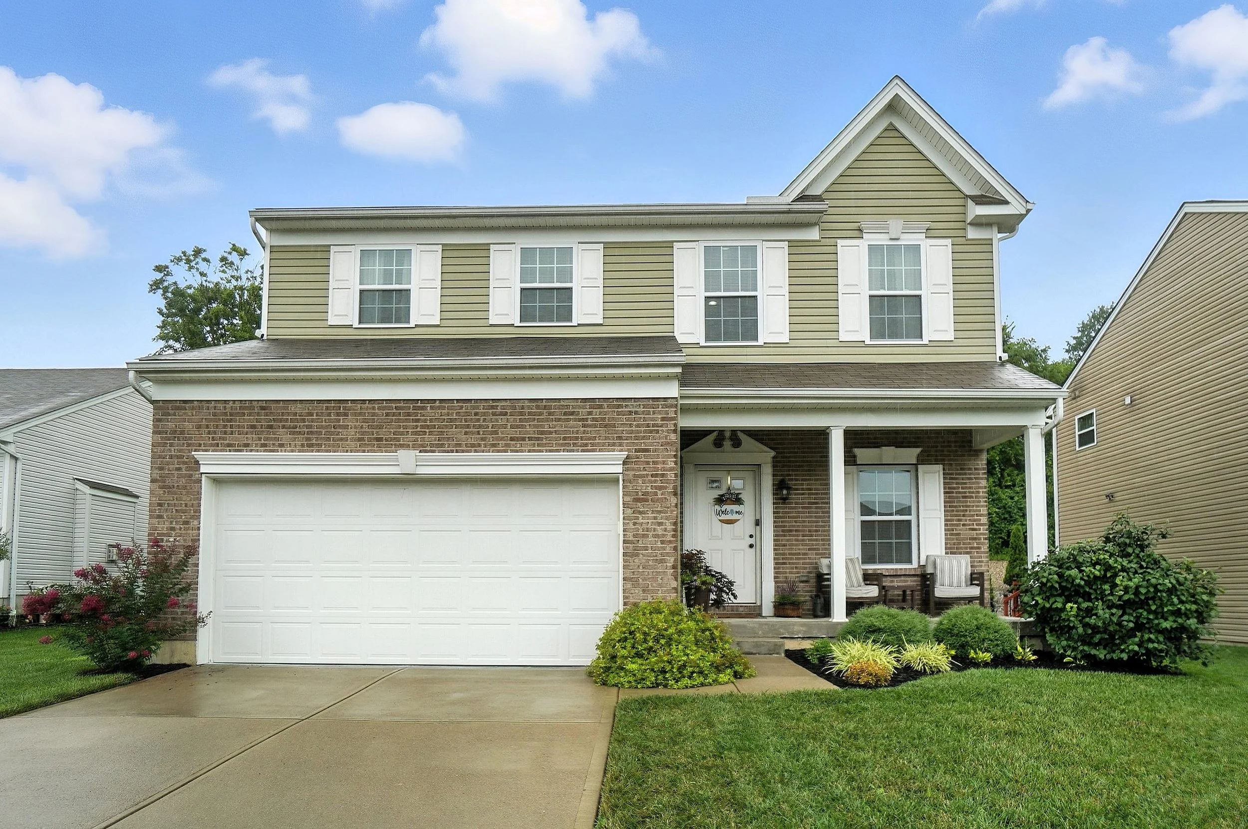 Two-story house with brick and yellow siding exterior, white garage door, front porch with seating, and landscaped front yard.