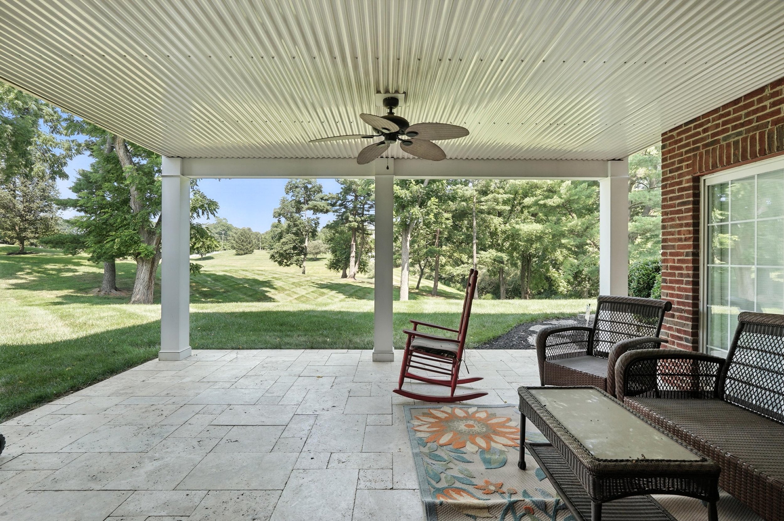 Covered patio with wicker chairs, a rocking chair, and a floral rug overlooking a green yard with trees and a lawn.