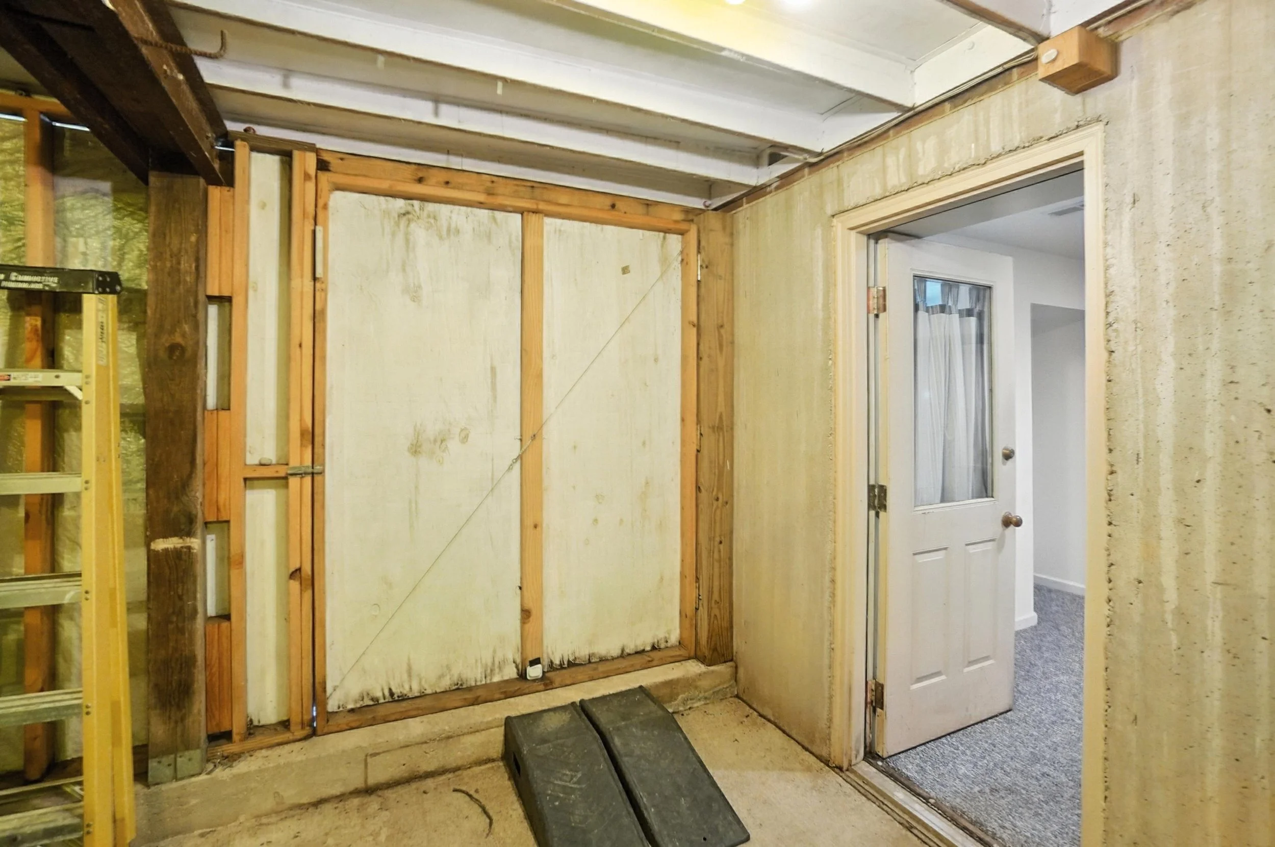 Interior view of a partially renovated room with an open door leading to another room with white walls and gray carpet. Visible construction materials include a ladder and wooden framing on the wall, with a strip of black material on the floor.