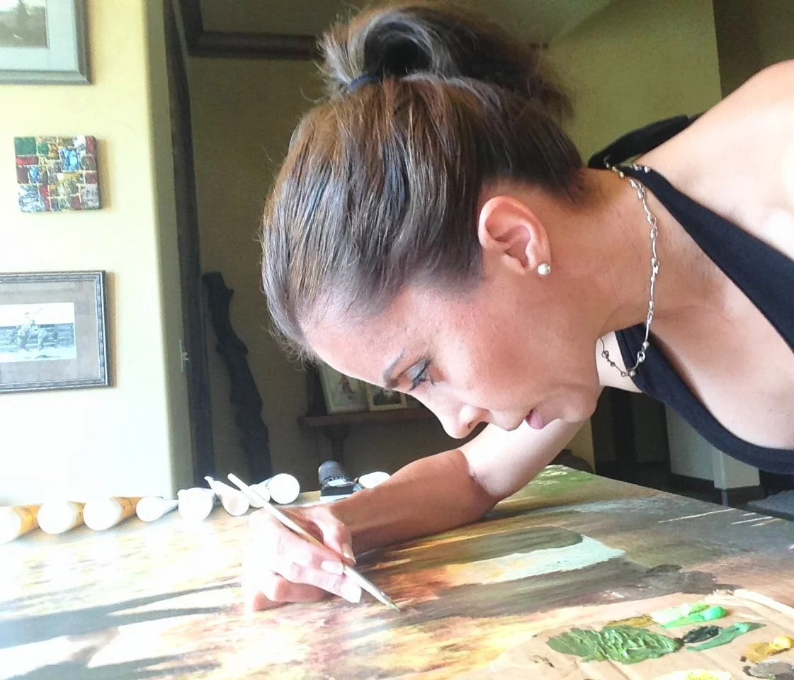 A woman with a ponytail, earrings, and a necklace is painting on a wooden surface with a small brush.