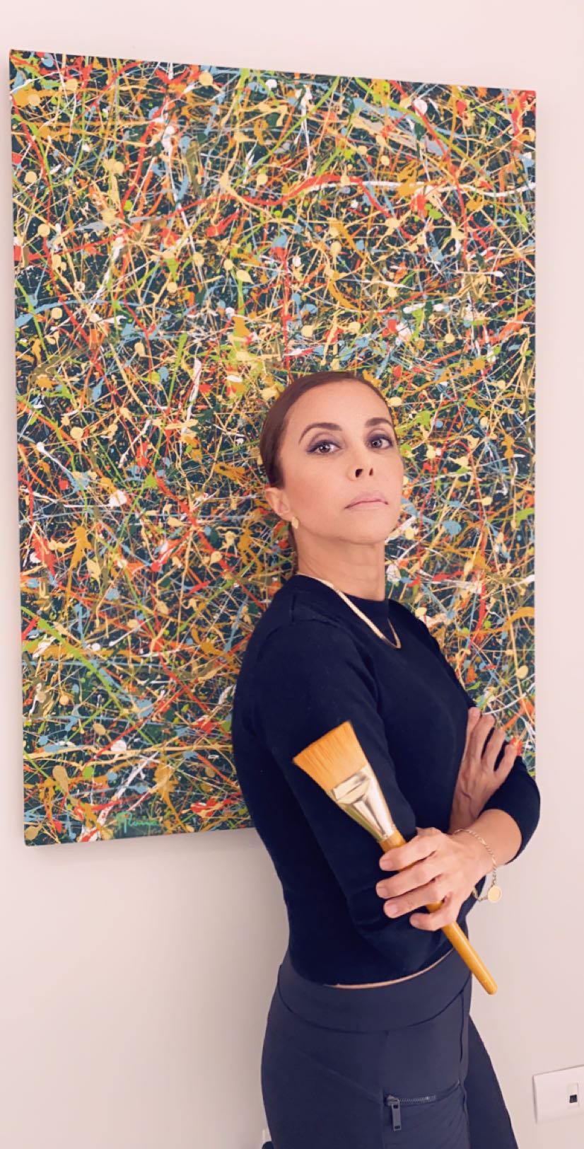 A woman with makeup and earrings poses with arms crossed, holding a large paintbrush, standing in front of an abstract splatter painting.