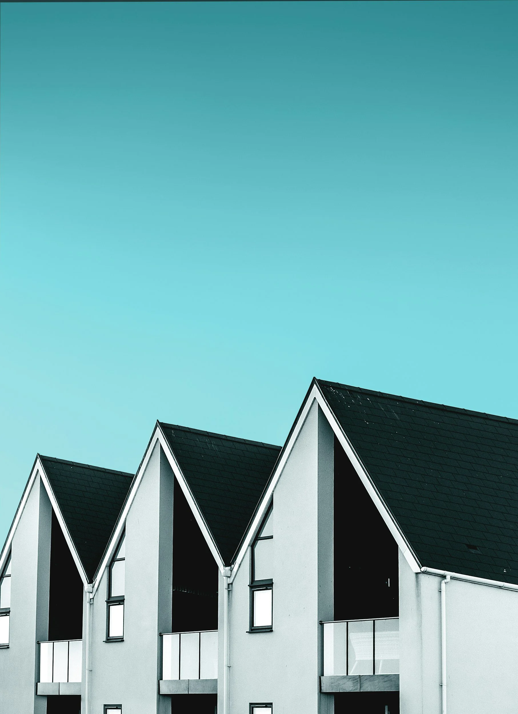 Modern white residential building with black tiled triangular roofs, glass balcony railings, and large windows under a clear blue sky.