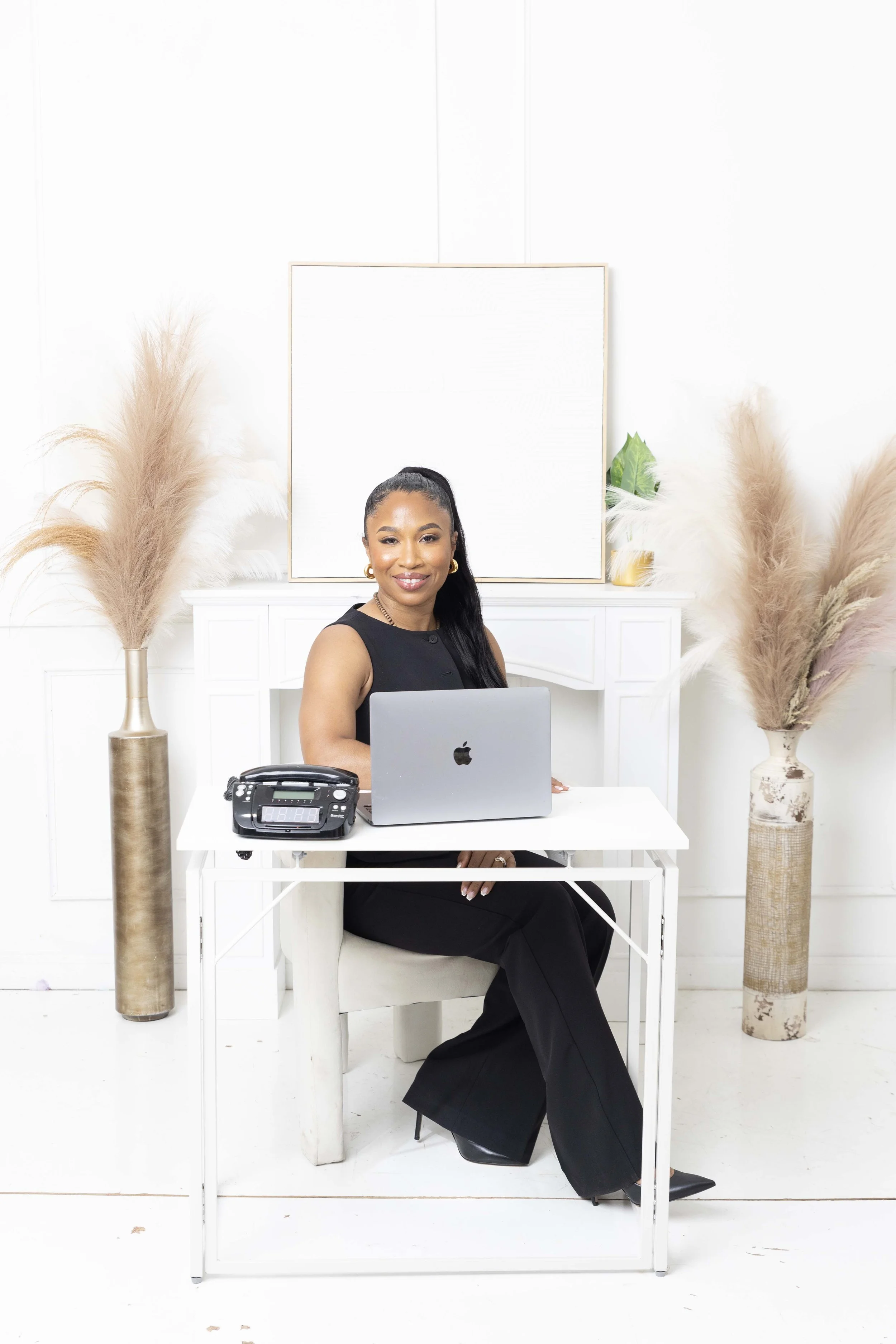 A woman with black hair in a high ponytail, wearing a sleeveless black top, sitting at a white desk with a laptop and office phone in a room decorated with large pampas grass arrangements and a blank white art canvas in the background.