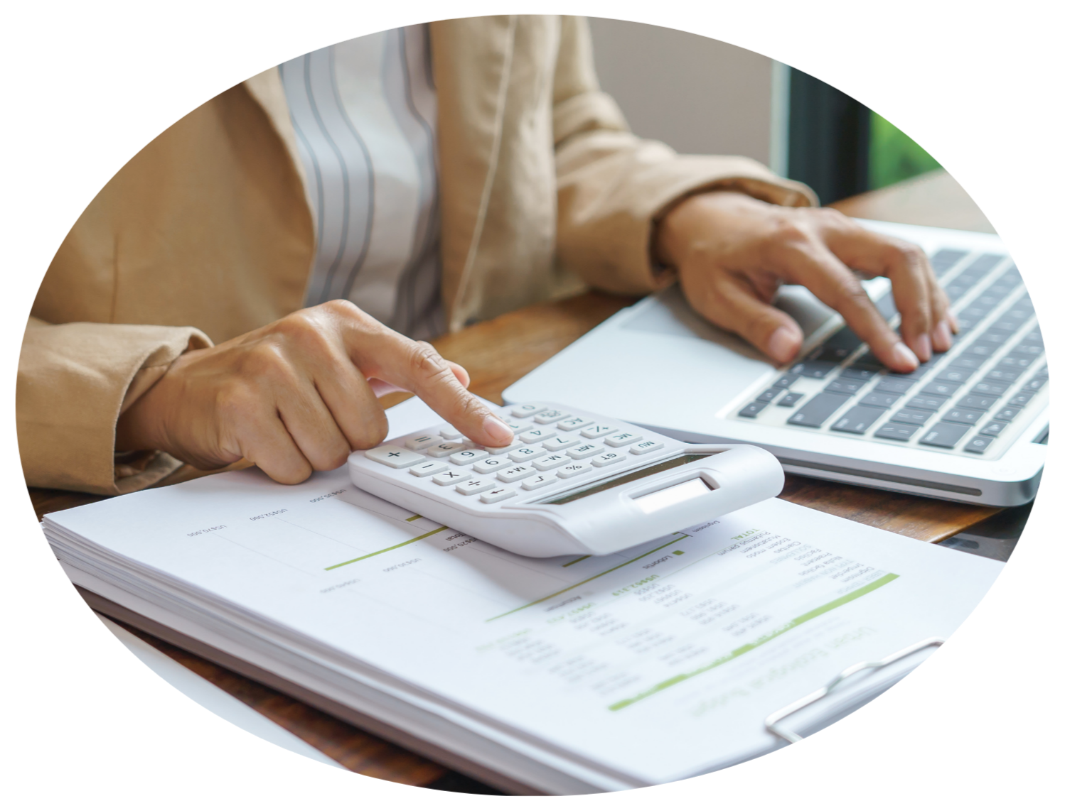 A person using a calculator and a laptop on a wooden desk, with a stack of papers and documents nearby.