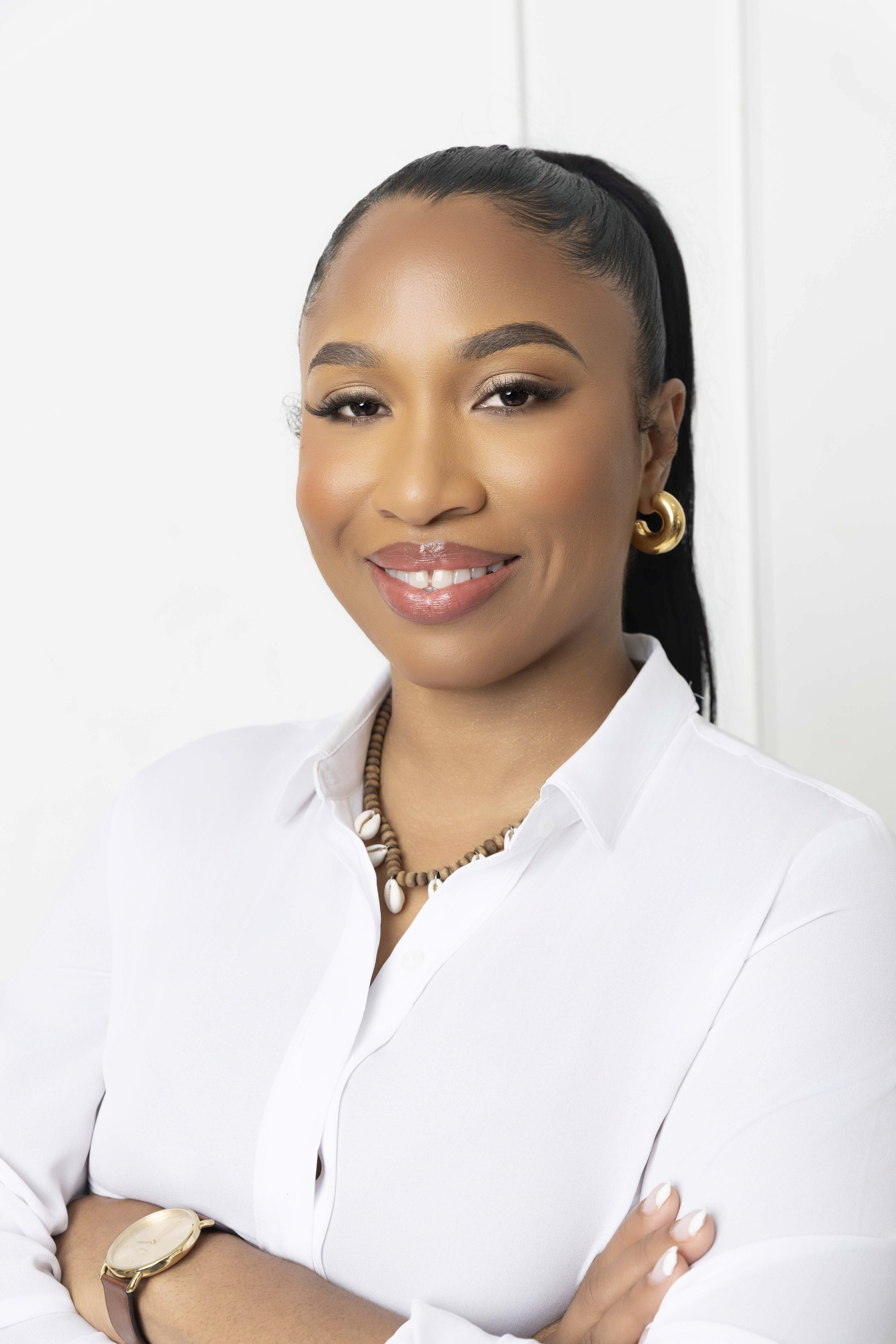 A woman with dark hair in a high ponytail, wearing a white collared shirt, gold hoop earrings, a necklace with shells, and a wristwatch, smiling confidently in front of a white background.