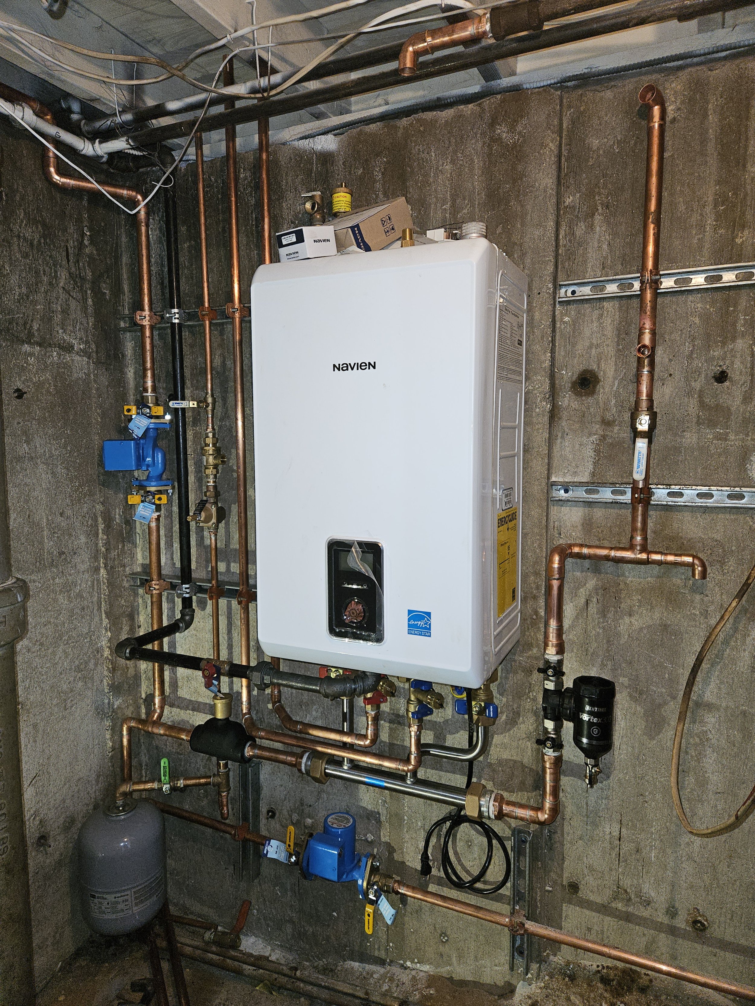 A plumbing and water heating system installed on a concrete wall, featuring copper pipes, a Navien water heater, and various valves and filters.