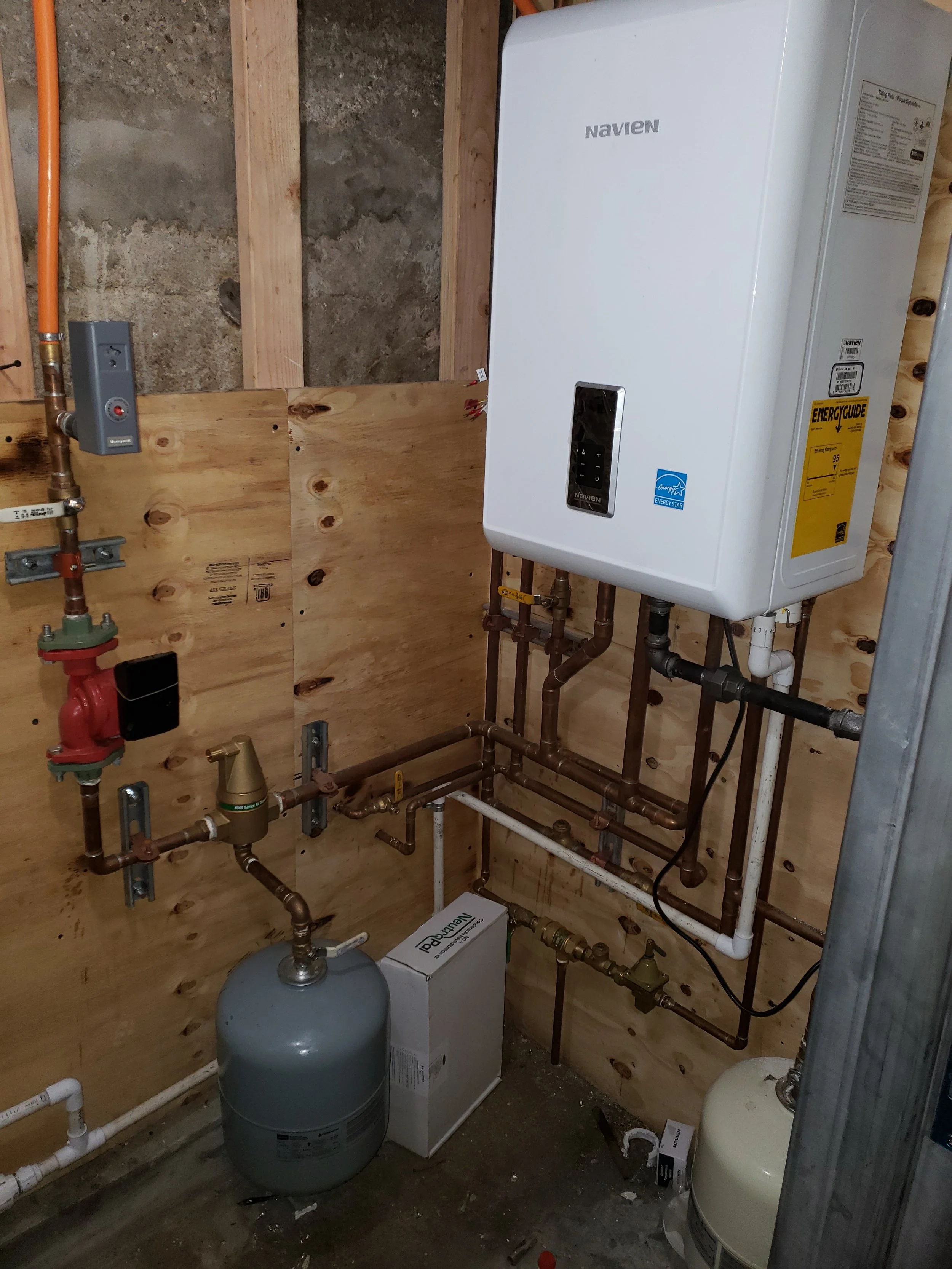 A utility room with exposed wooden framing, plumbing, and two water heaters, one white Navien tankless heater and one smaller gray tank.