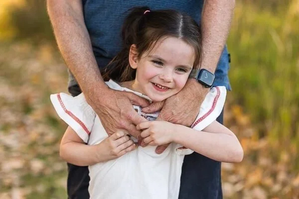 Little girl leans against her father & smiles as he wraps his arms around her shoulders