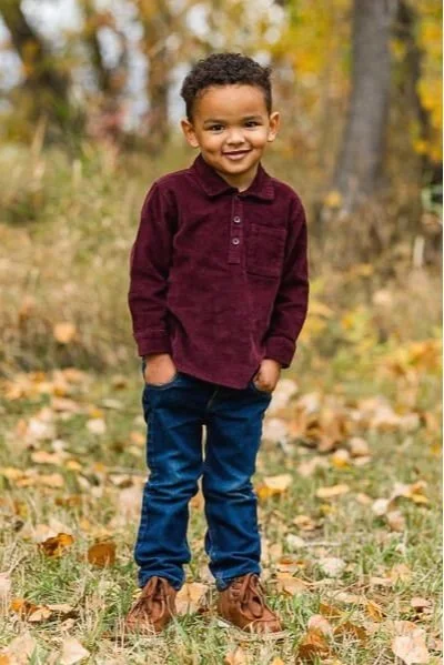 Little boy in burgundy shirt standing with hands in pockets
